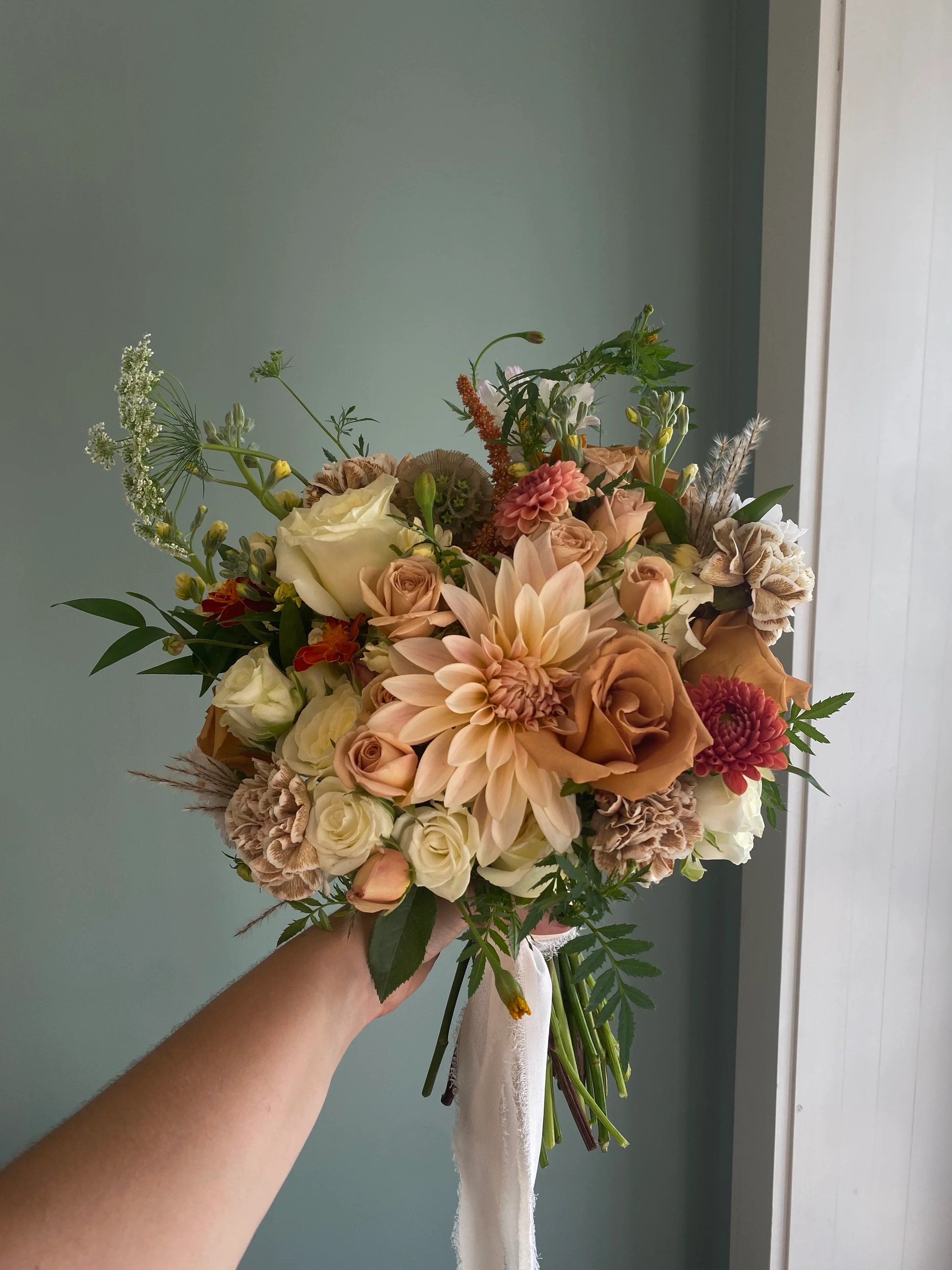 Hand holding a bouquet of mixed flowers including roses, dahlias, and greenery against a light green wall