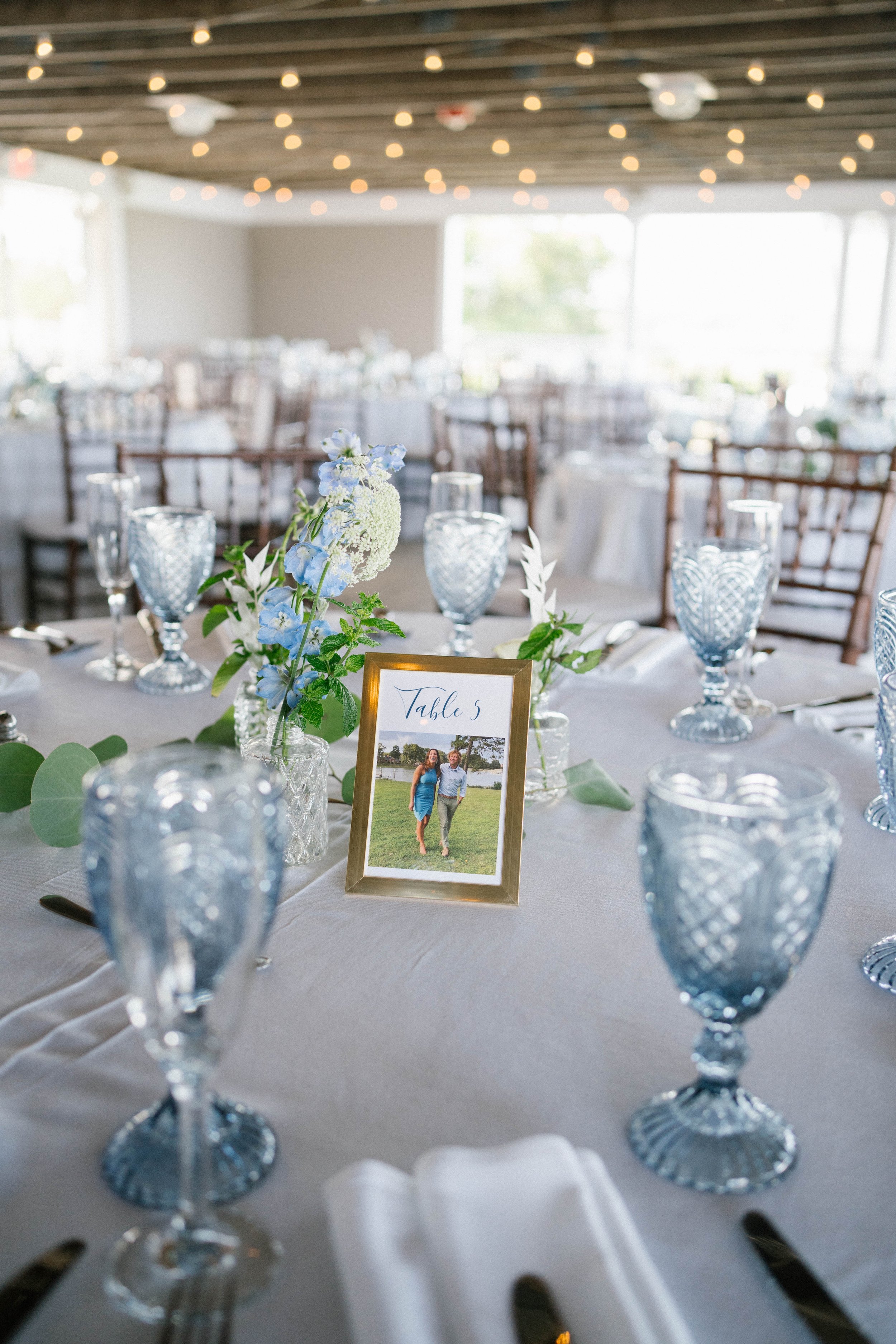 Wedding reception table decorated with blue glassware, floral centerpiece, and a framed photo indicating "Table 5."