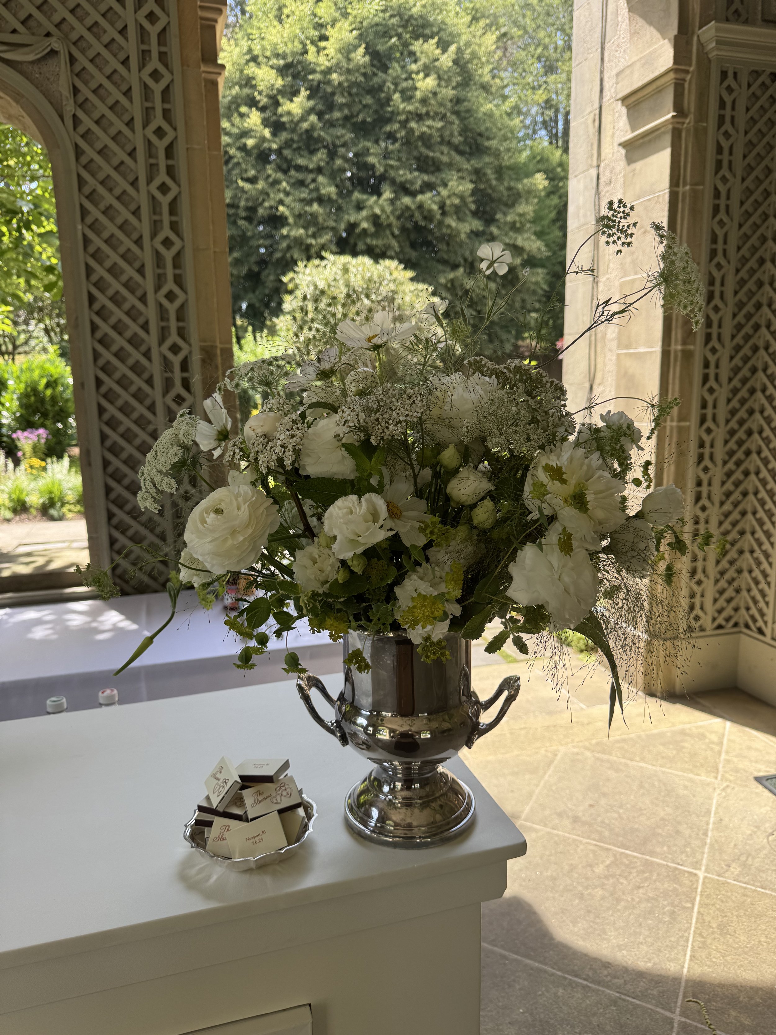 Large silver vase with white floral arrangement on a white table inside a building, with an open door and green garden outside in the background.