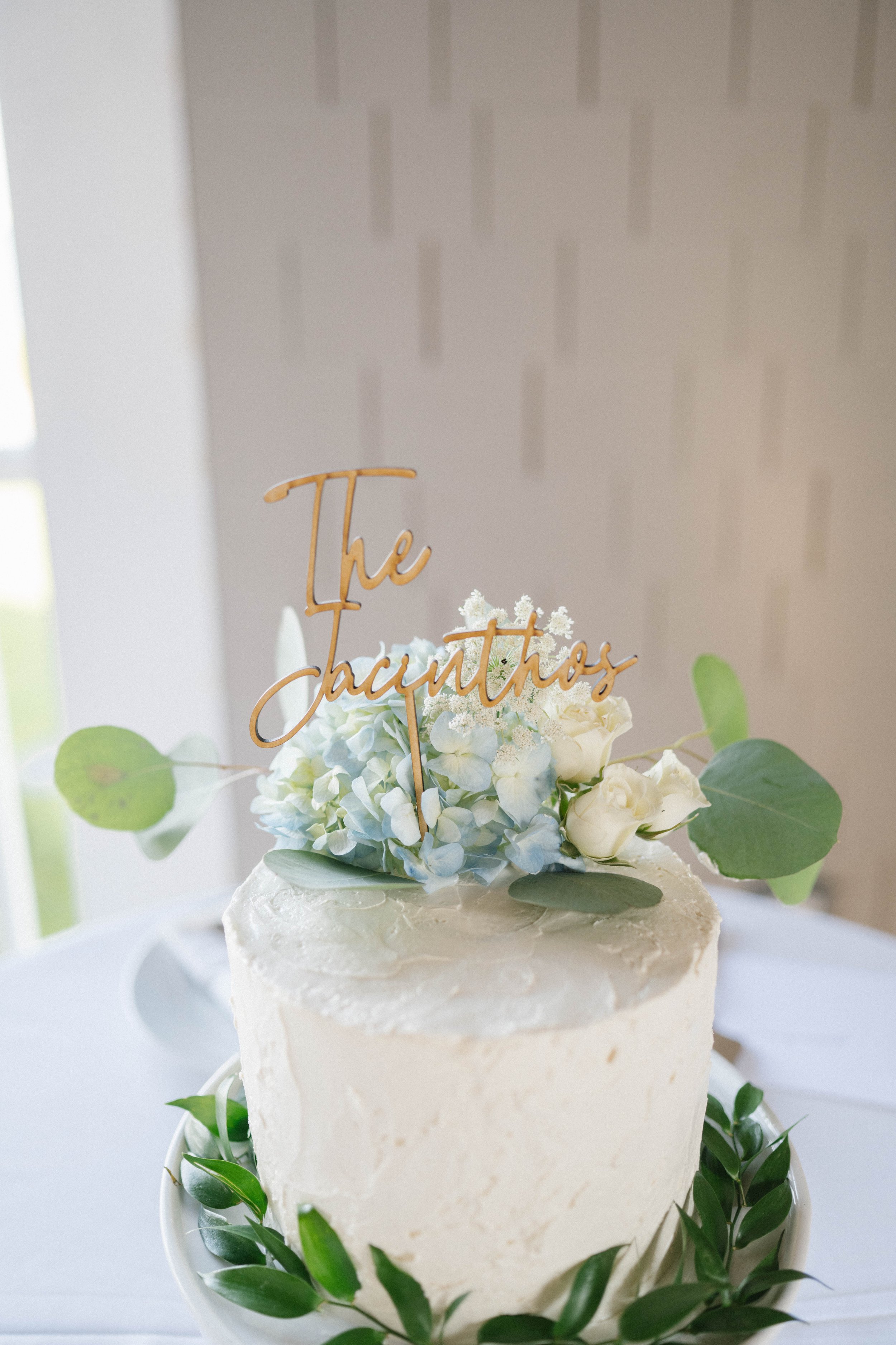 A white frosted wedding cake with green leaves on the base, topped with white and pale blue flowers, and a wooden cake topper that reads 'The Jacintos'.