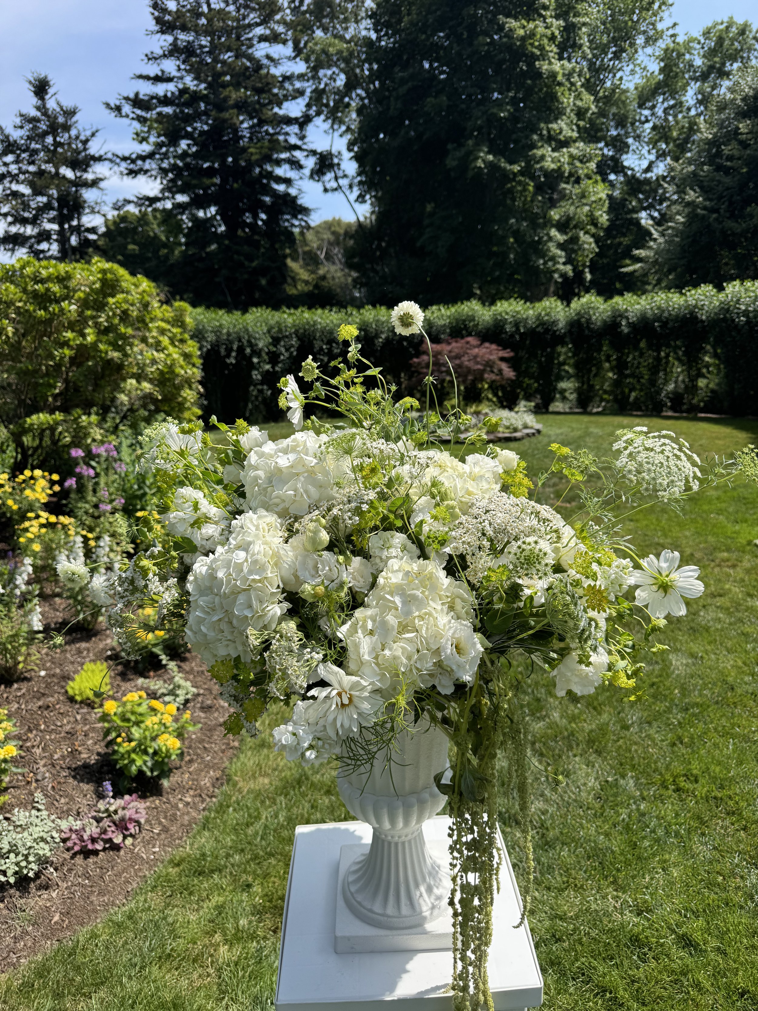 White flower arrangement in a white vase on a white pedestal in a garden with green grass, bushes, and trees.