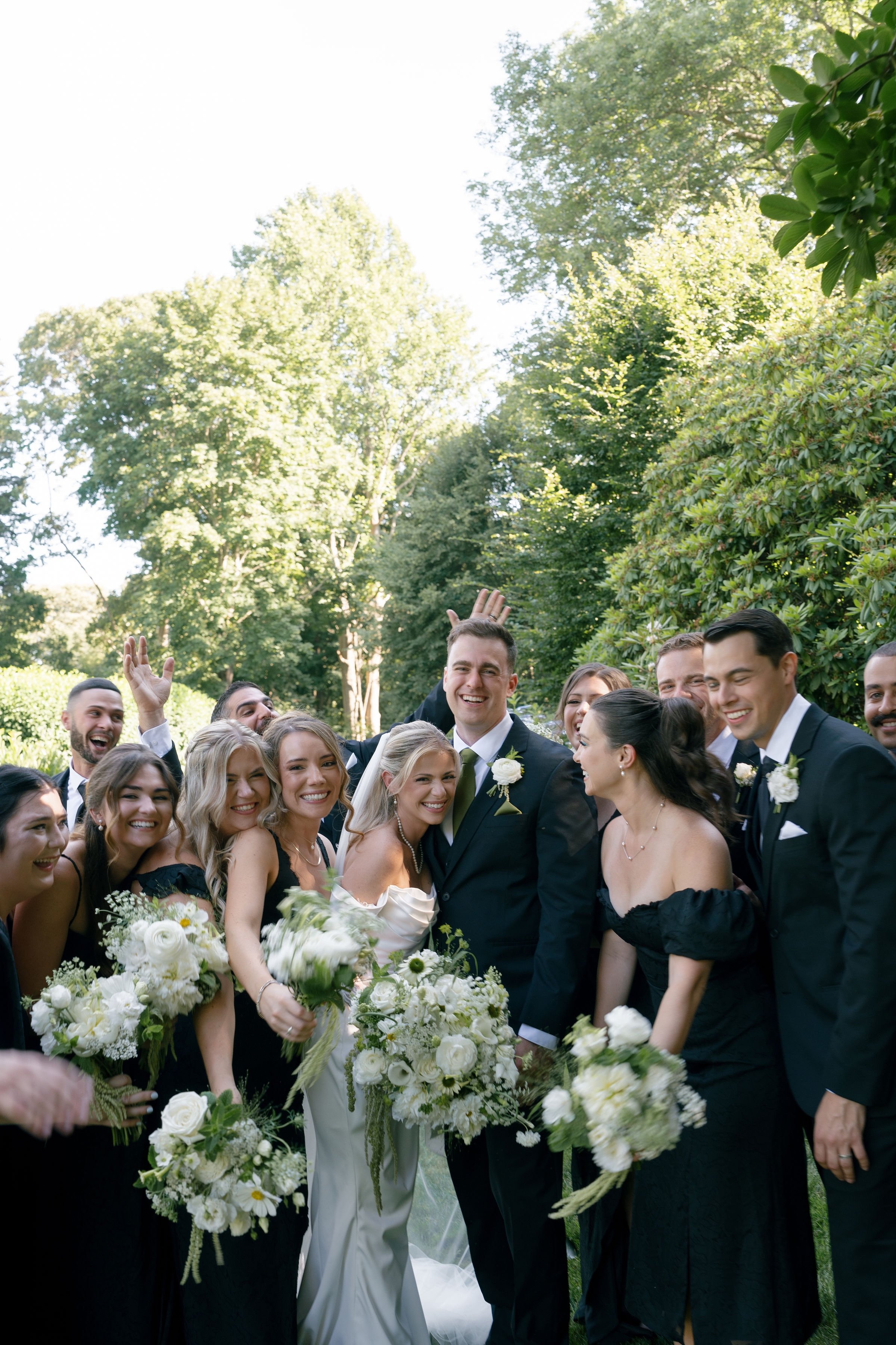 A wedding party outdoors with the bride and groom in the center surrounded by friends and family holding bouquets of white flowers, on a sunny day with green trees in the background.