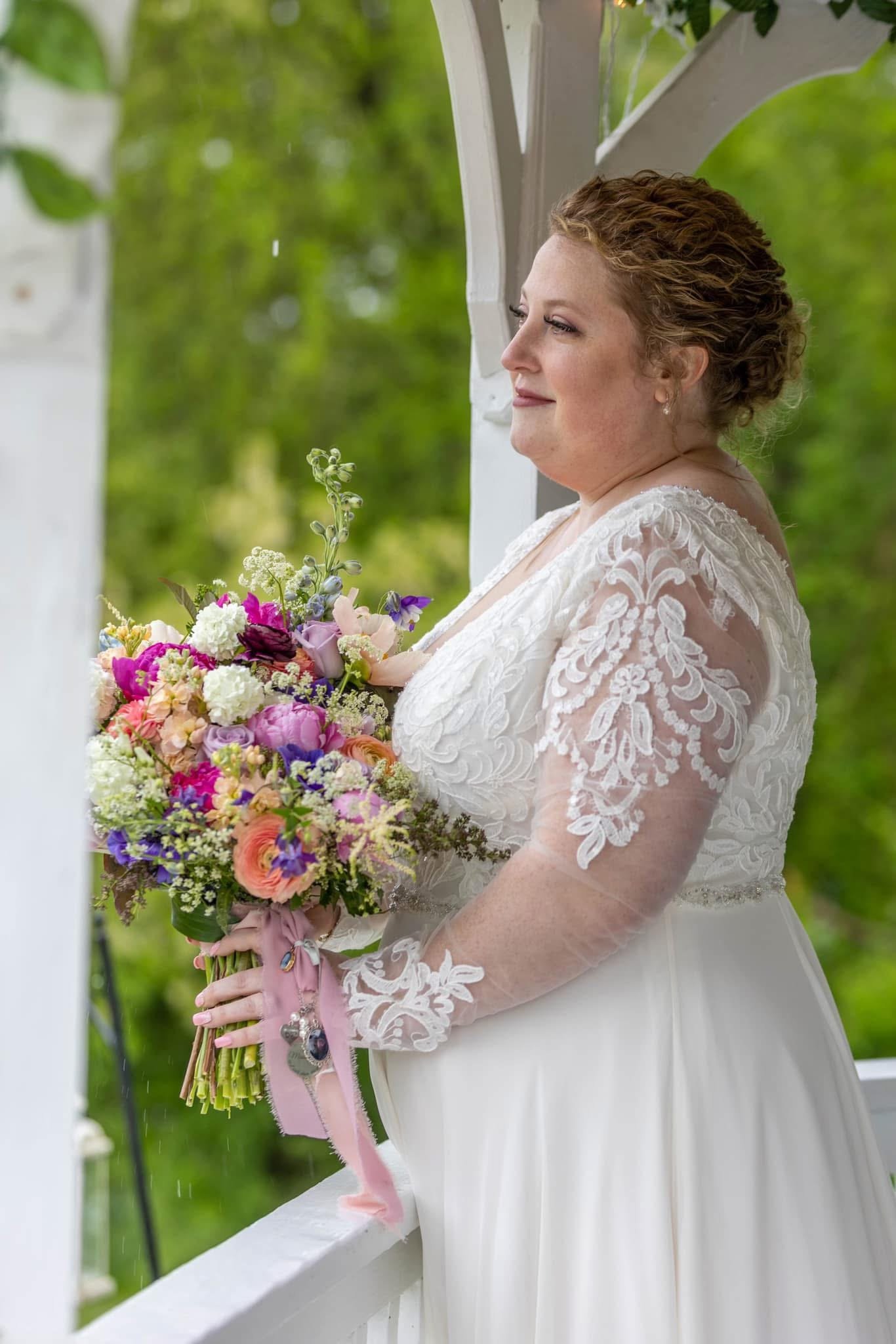 A bride holding a colorful bouquet of flowers, dressed in a white lace wedding gown, standing on a porch with green foliage in the background.