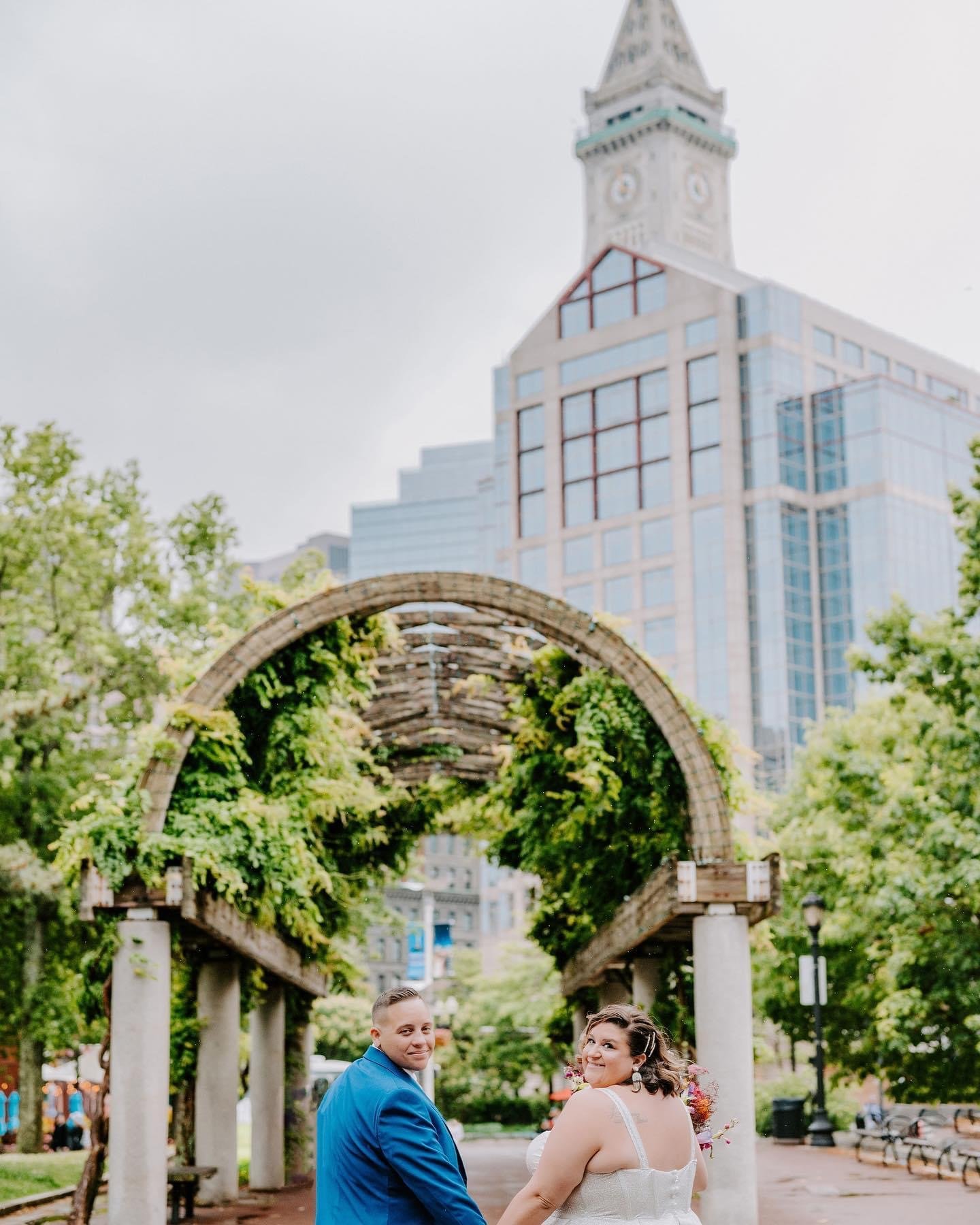 A couple in wedding attire standing under a wooden arch covered with greenery in a city park with skyscrapers in the background.