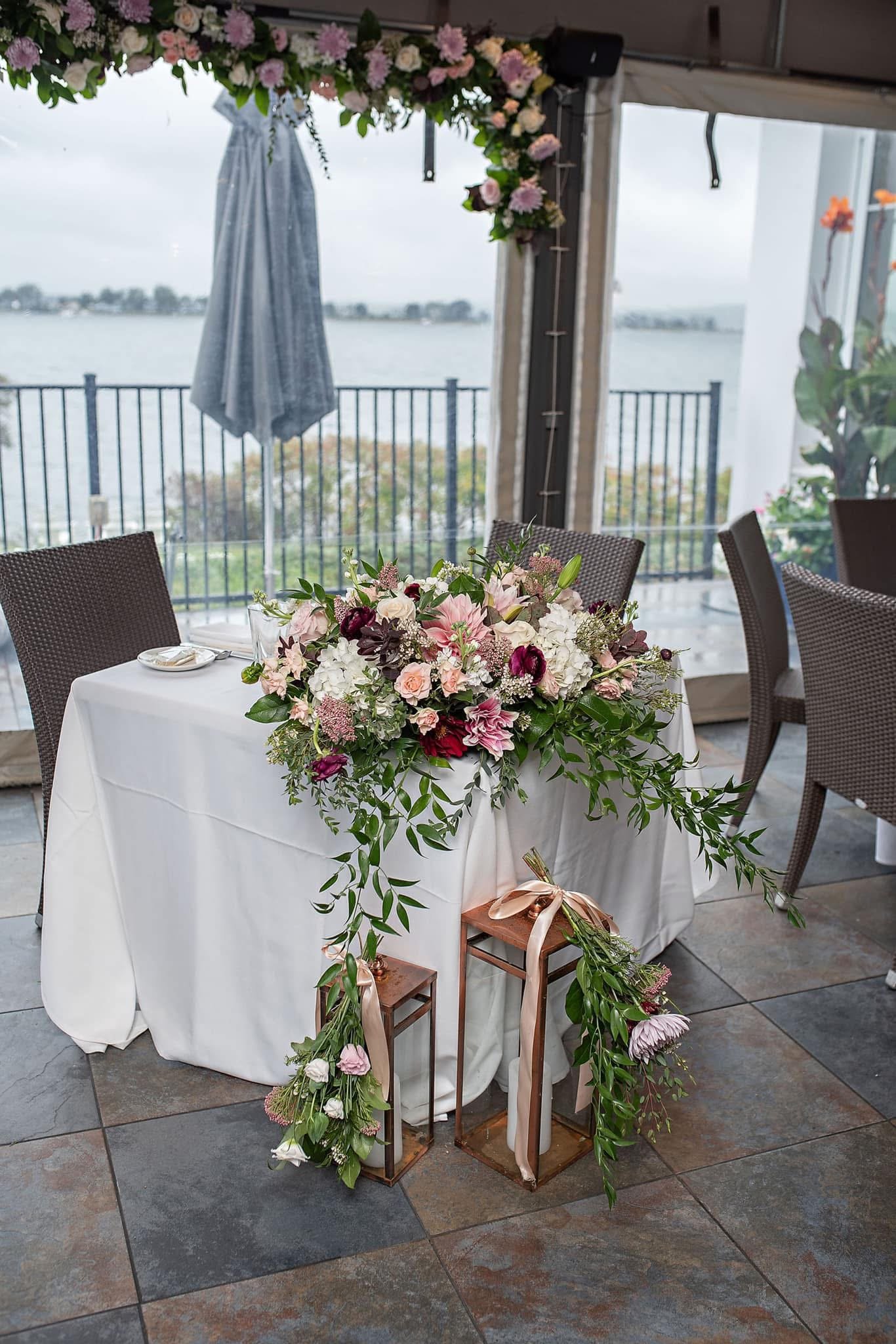 A table decorated with a large floral centerpiece of pink, white, and purple flowers, set in front of a lakeside window with an umbrella and outdoor scenery in the background.