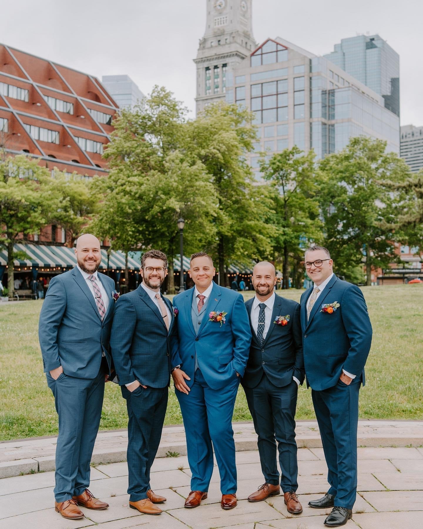 Five men in suits standing together outdoors in a park, smiling, with tall buildings and trees in the background.