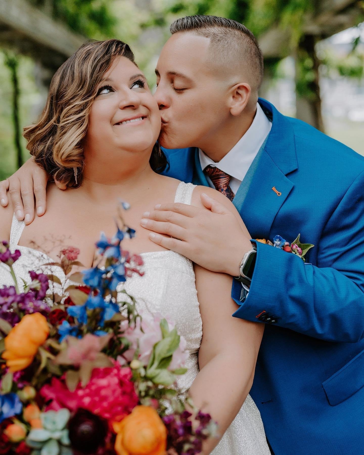 A man in a blue suit kisses a woman in a white dress on the cheek at an outdoor wedding, with colorful flowers in the foreground and green trees in the background.