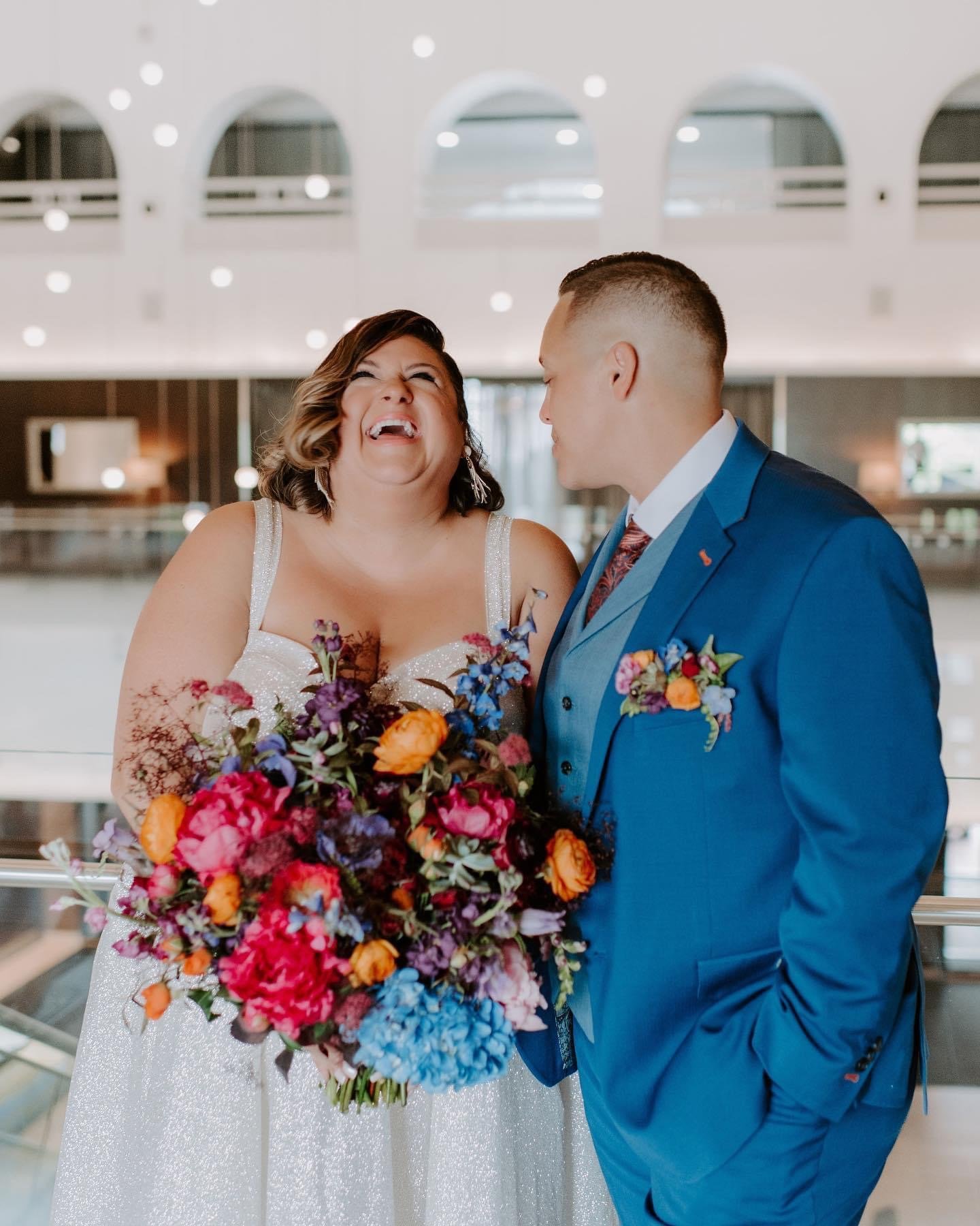 A woman in a white dress holding a large bouquet of colorful flowers, laughing happily, standing next to a man in a blue suit, smiling, in an elegant indoor setting with arched windows.