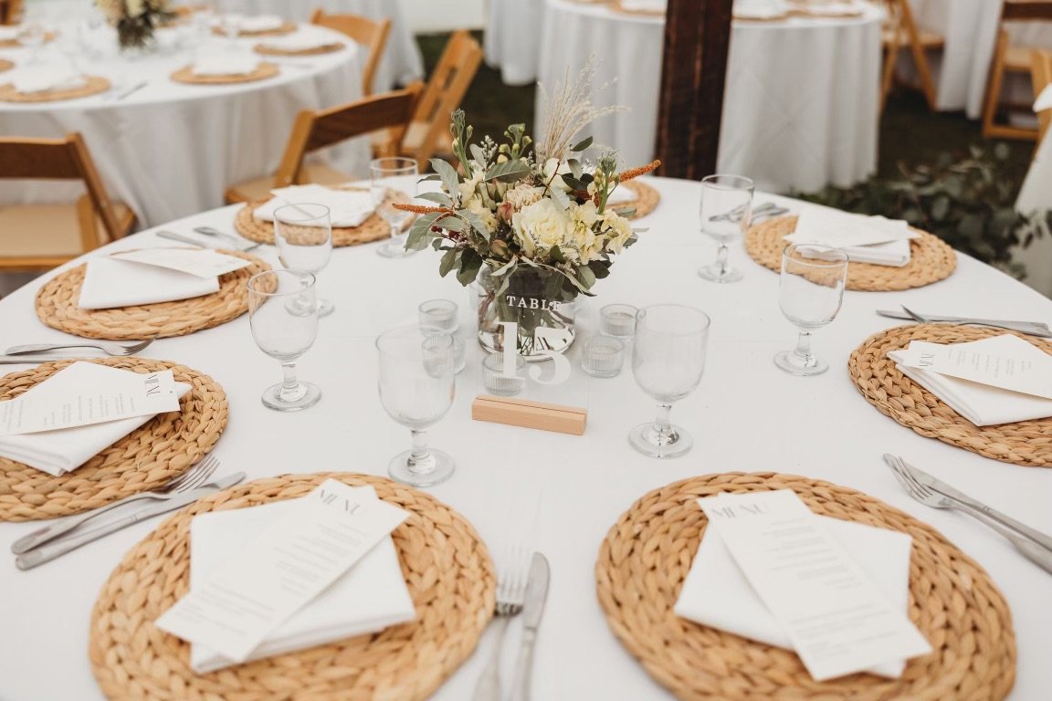 Round table set for a wedding or event with a white tablecloth, woven placemats, white napkins, utensils, clear glasses, a floral centerpiece, and a table number sign.