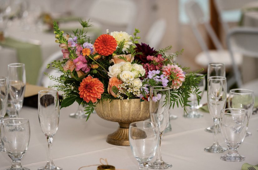 A colorful floral centerpiece with pink, orange, white, and purple flowers in a gold bowl on a decorated table with empty glassware.