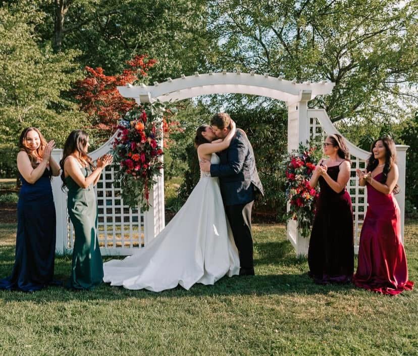 A newly married couple sharing a kiss under a white garden arch decorated with red and pink flowers, surrounded by four bridesmaids clapping and smiling in an outdoor setting with green trees in the background.