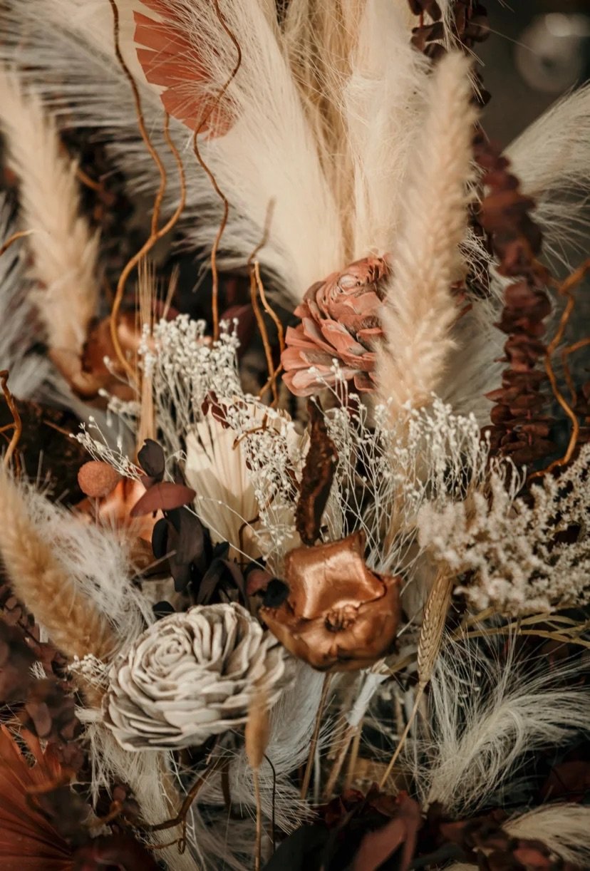 A close-up of a floral arrangement with dried and preserved flowers, including pampas grass, roses, and other botanical elements in warm, earthy tones.