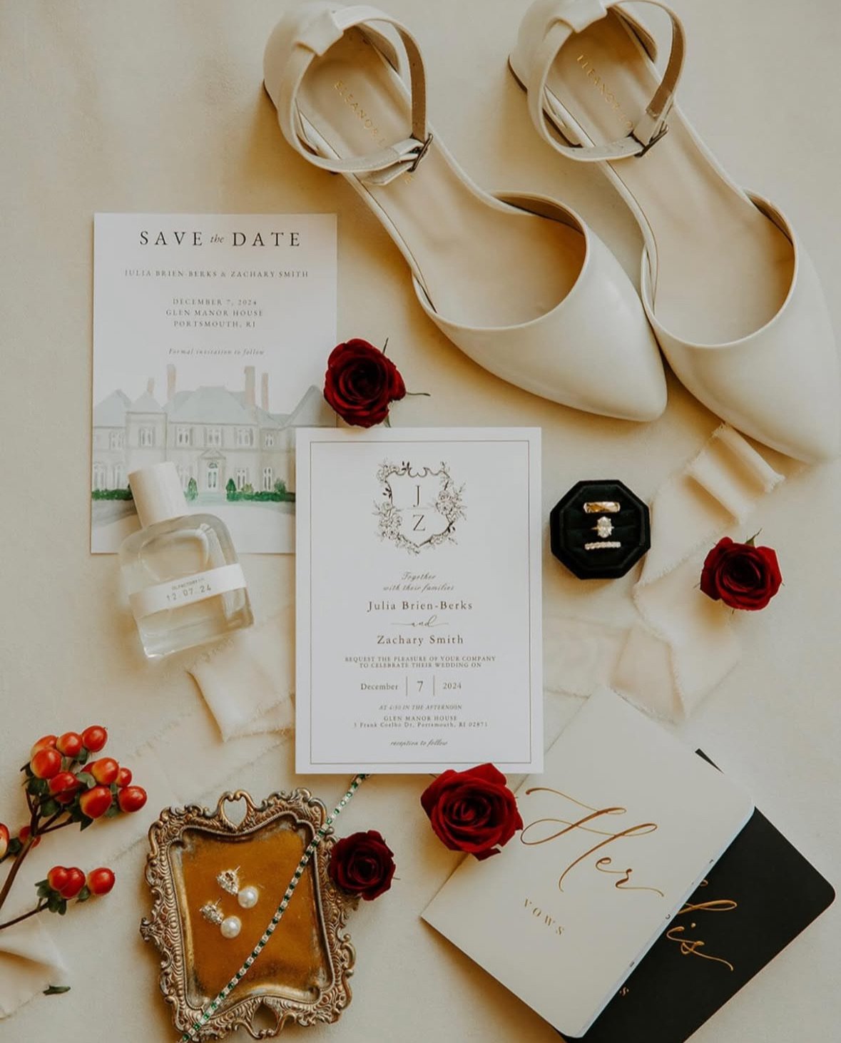 Wedding invitation suite with white heels, red roses, jewelry, candles, and decorative items on a cream surface.