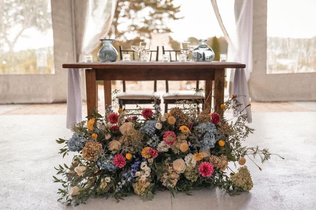 A dining table set with glassware and vases, with a large colorful flower arrangement in front of it. The setting appears to be indoors with curtains and windows in the background.