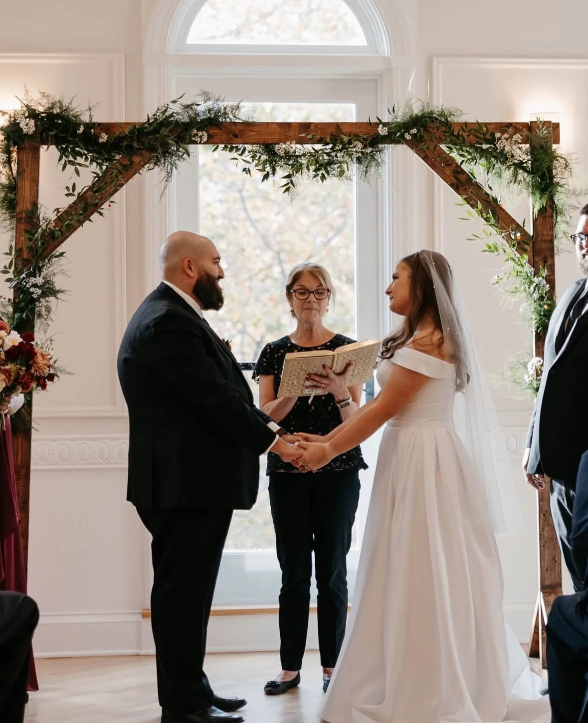 A bride and groom holding hands during their wedding ceremony inside a decorated room with a wooden arch and natural light.