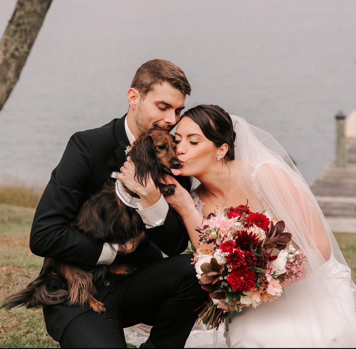 A bride and groom are kneeling outdoors by a body of water, holding a dog between them. They are affectionately kissing and hugging the dog, who is wearing a bowtie. The bride is holding a bouquet of red, pink, and white flowers. The groom is in a bl