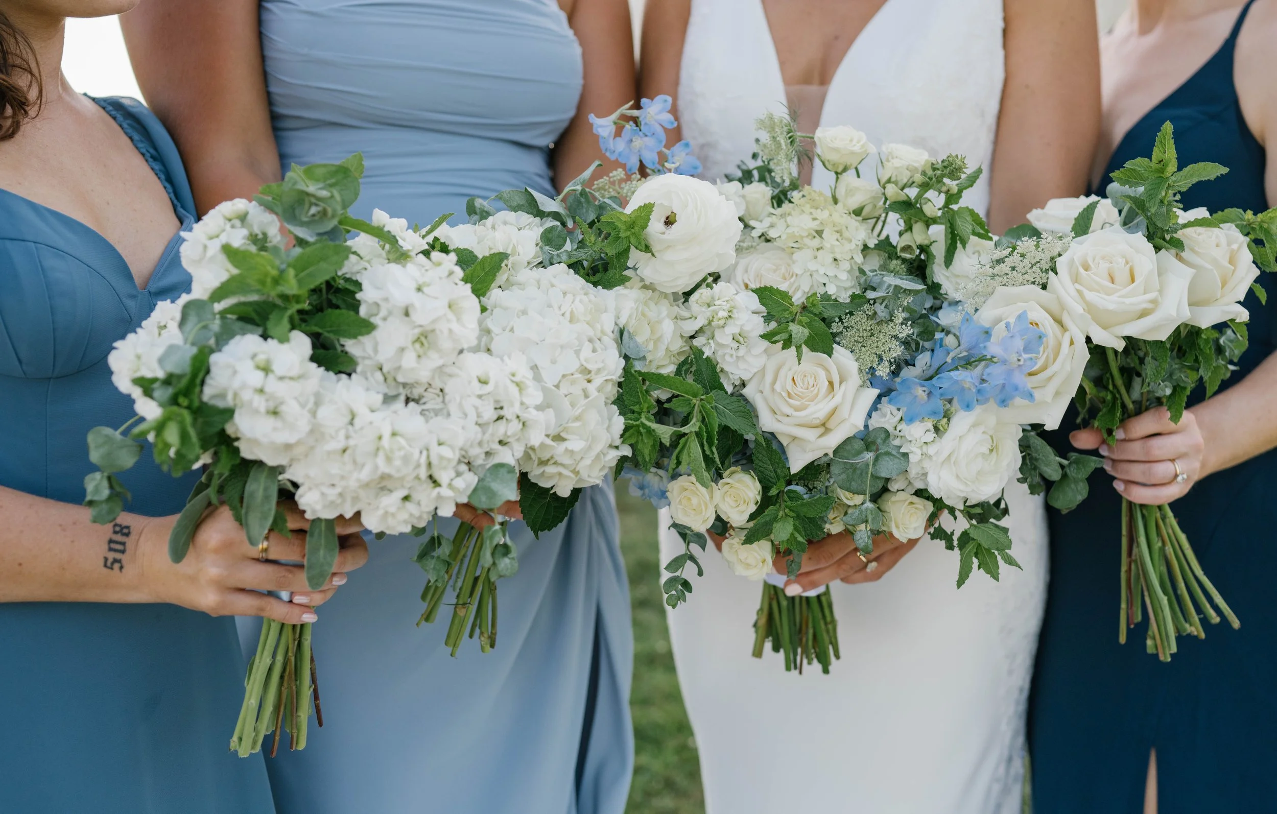 Close-up of three women holding bouquets of white and light blue flowers during a wedding, with the bride in a white dress in the center.