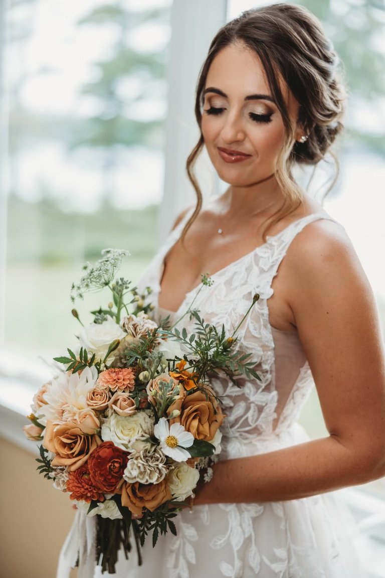 A bride with wavy brown hair styled in an updo, wearing a lace wedding gown, holding a colorful bouquet of flowers including roses, dahlias, and daisies, standing indoors with a blurred window background.