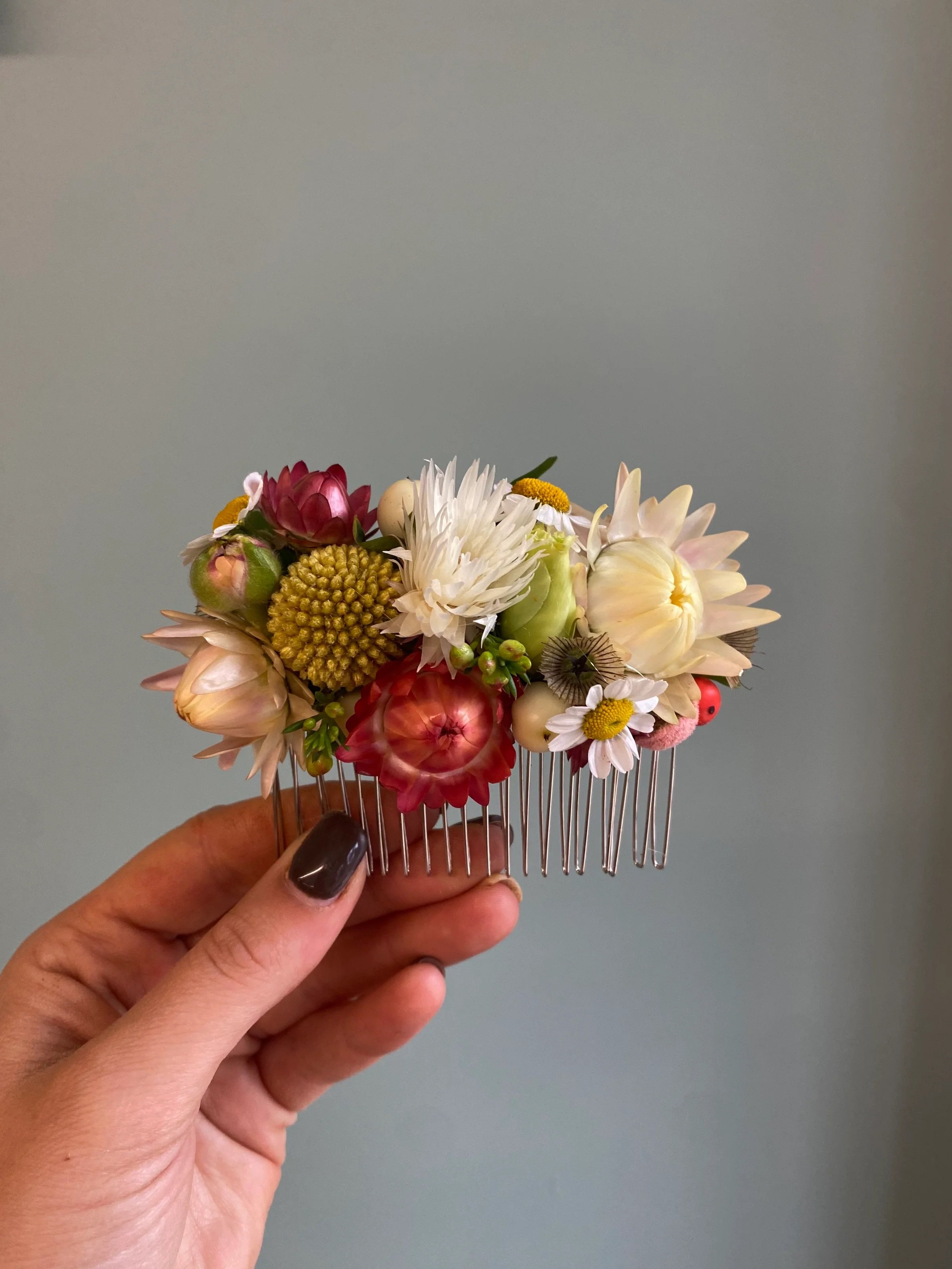 Hand holding a floral hair comb with various colorful flowers, including pink, white, yellow, and cream blooms, against a plain gray background.