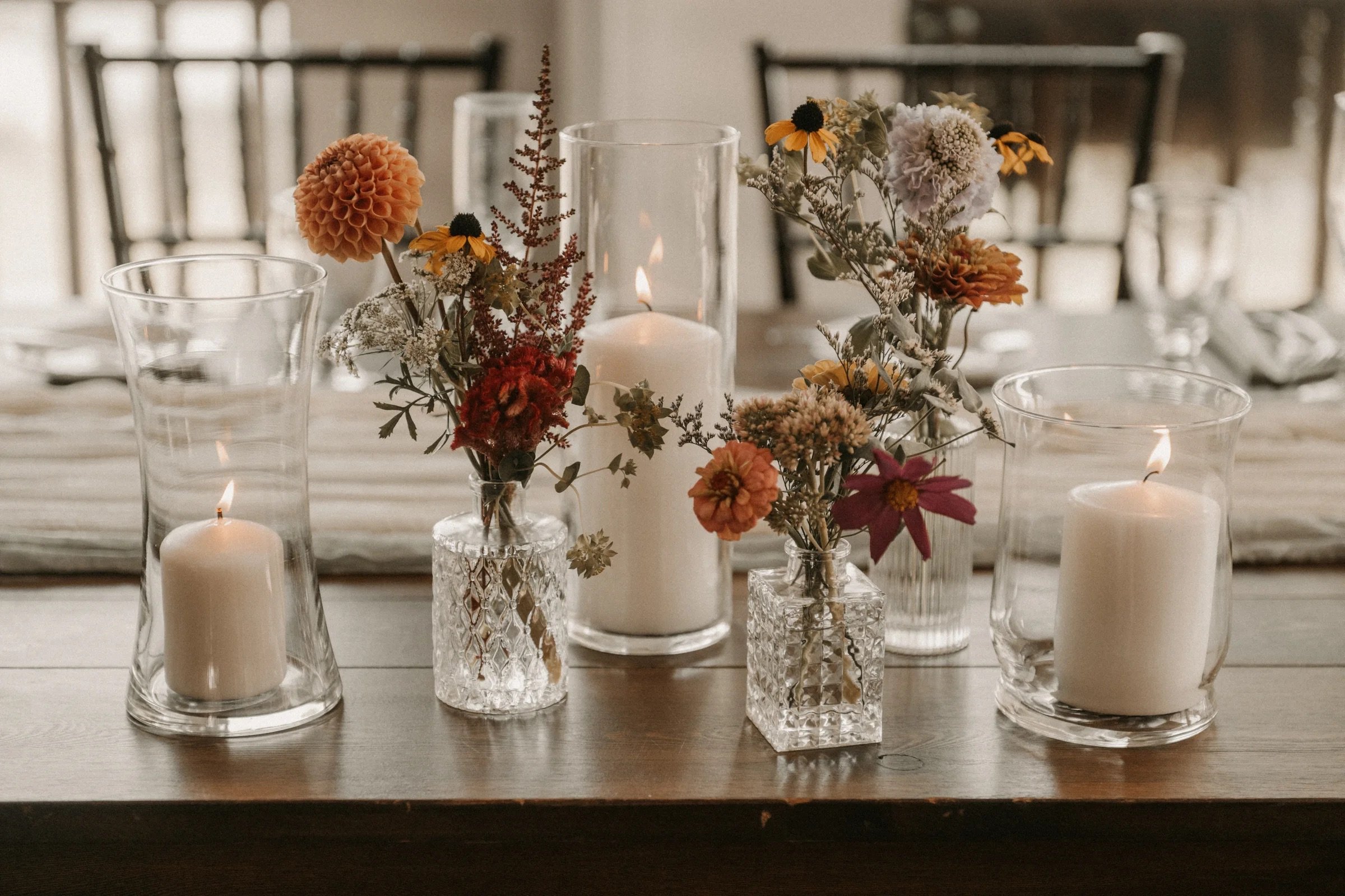 Table centerpiece with three glass vases holding various flowers, two white candles in glass holders, and a large white candle in a glass cylinder, on a wooden table.