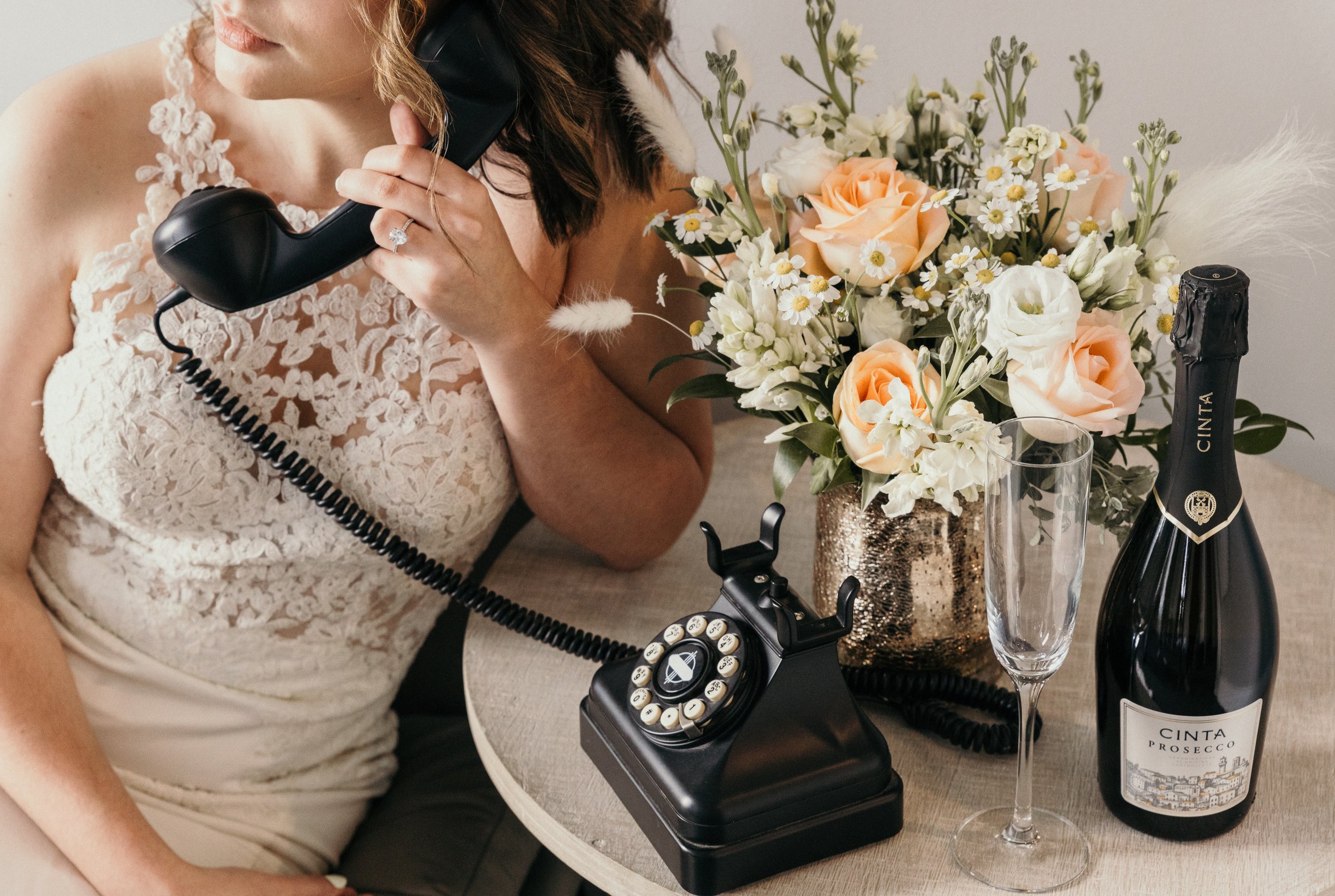 Woman in a lace dress talking on a vintage black rotary phone at a table with a bouquet of white and peach flowers, a bottle of Cinta Prosecco, and an empty champagne flute.