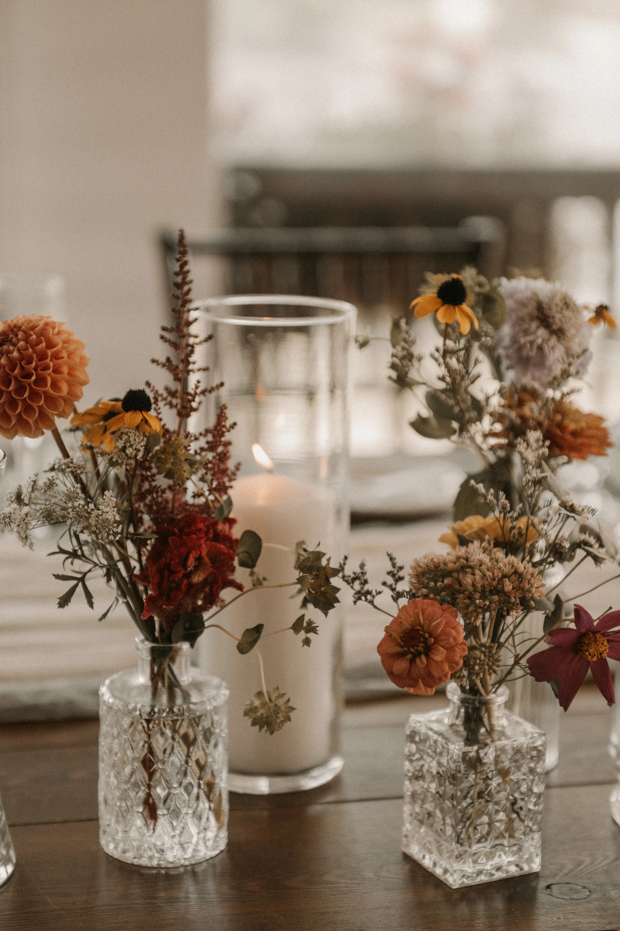A table centerpiece of mixed dried flowers in textured glass vases, with a white candle in a glass holder and a lit candle in the background.
