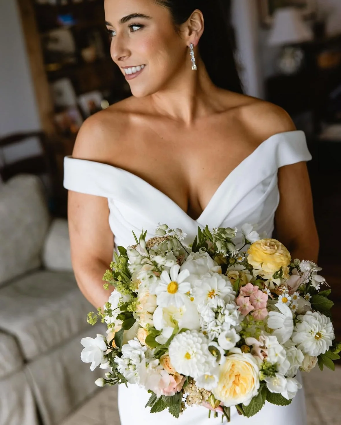 A bride in a white off-the-shoulder wedding dress holding a bouquet of white, yellow, and blush flowers, smiling and looking to her left.