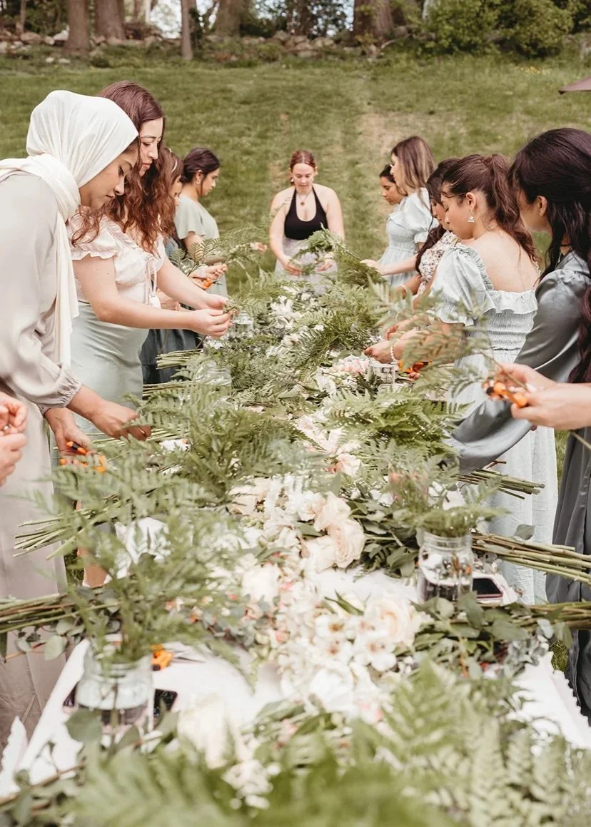 Women standing around a table outdoors making flower arrangements with greenery and flowers.