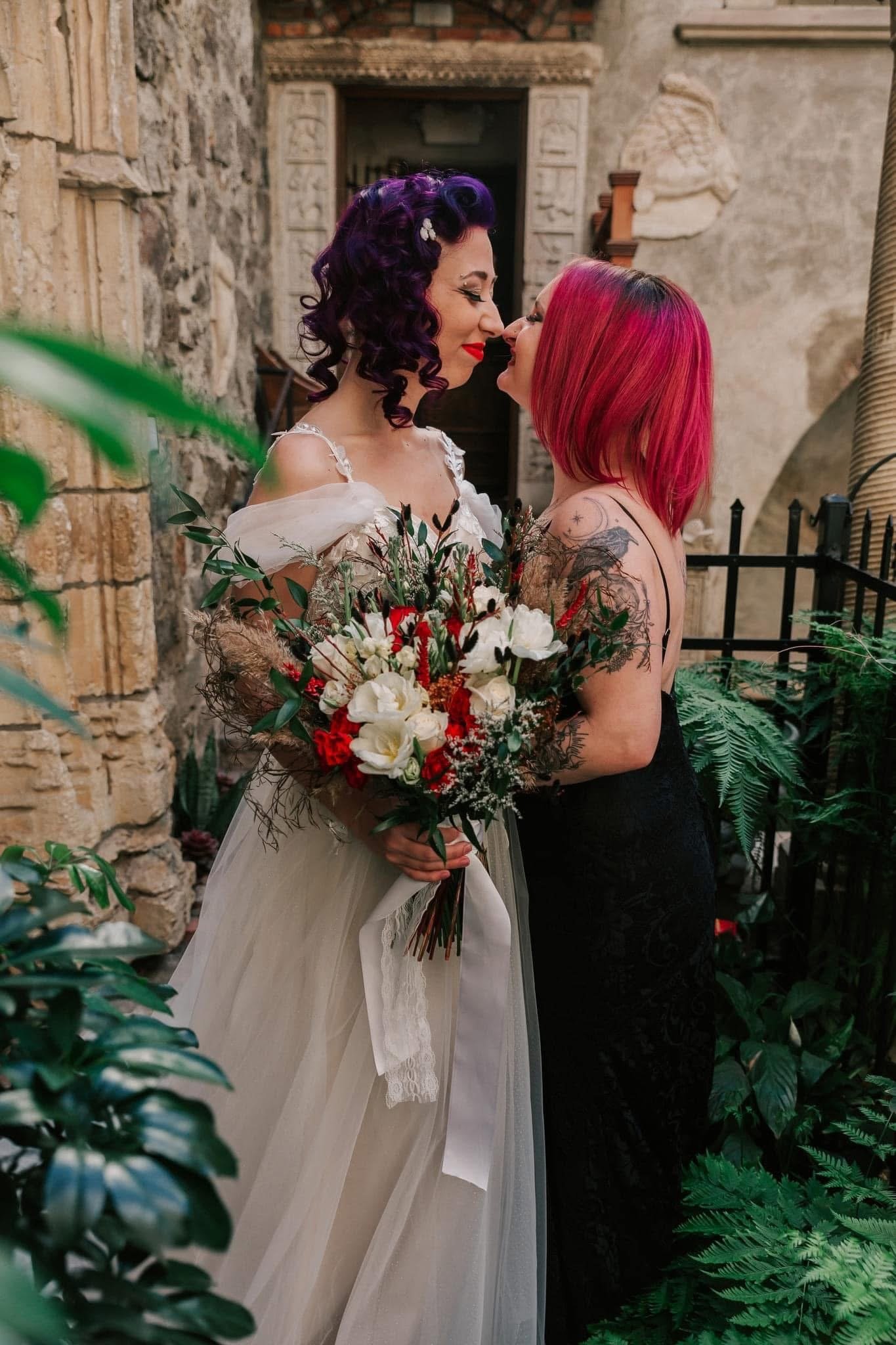 Two women, one in a wedding dress with dark purple curls holding a flower bouquet, and the other with bright pink hair in a black dress, are touching noses in an outdoor setting with stone and brick walls and green plants.