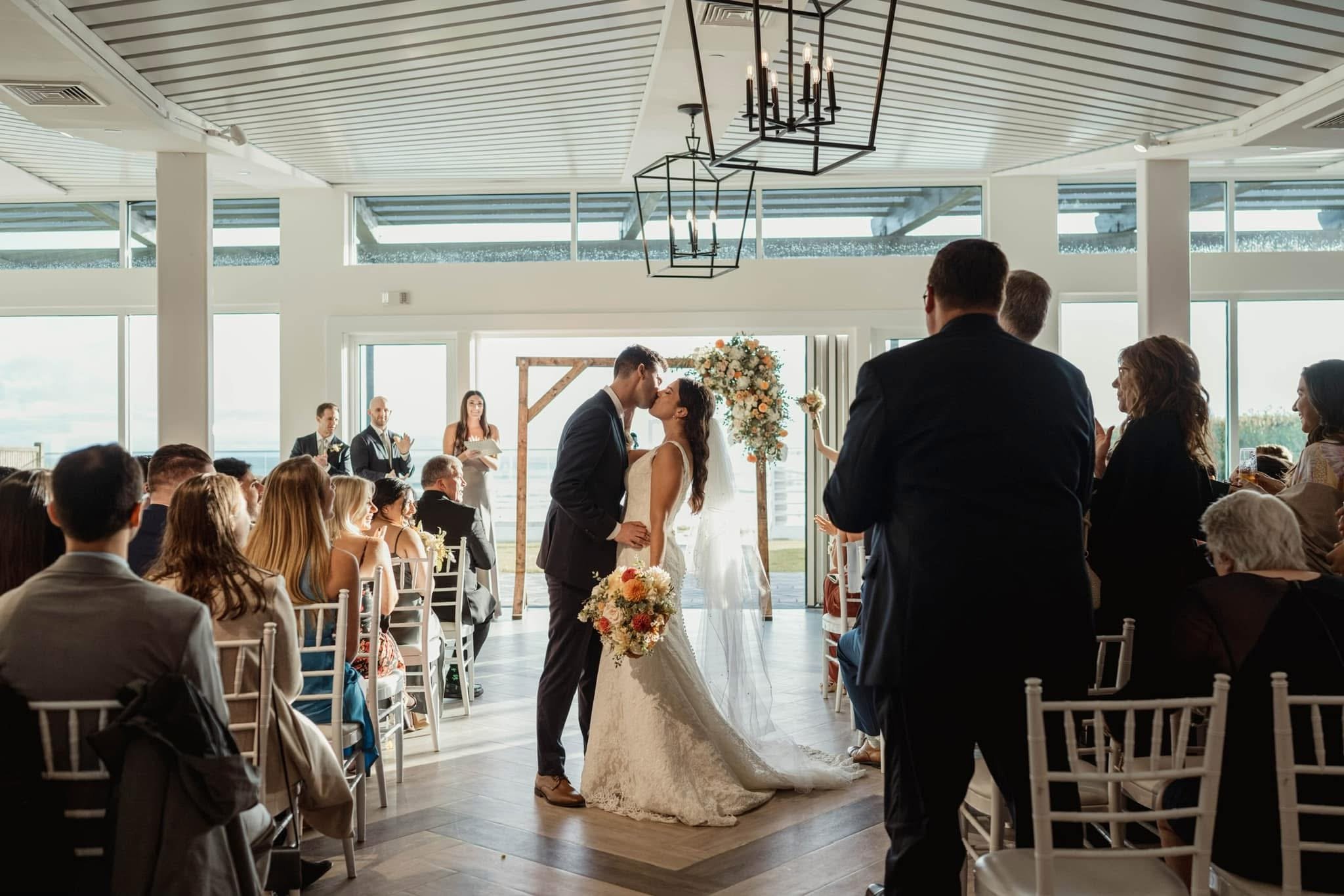 A bride and groom kiss during their wedding ceremony in a bright, modern indoor venue with large windows and an ocean view.