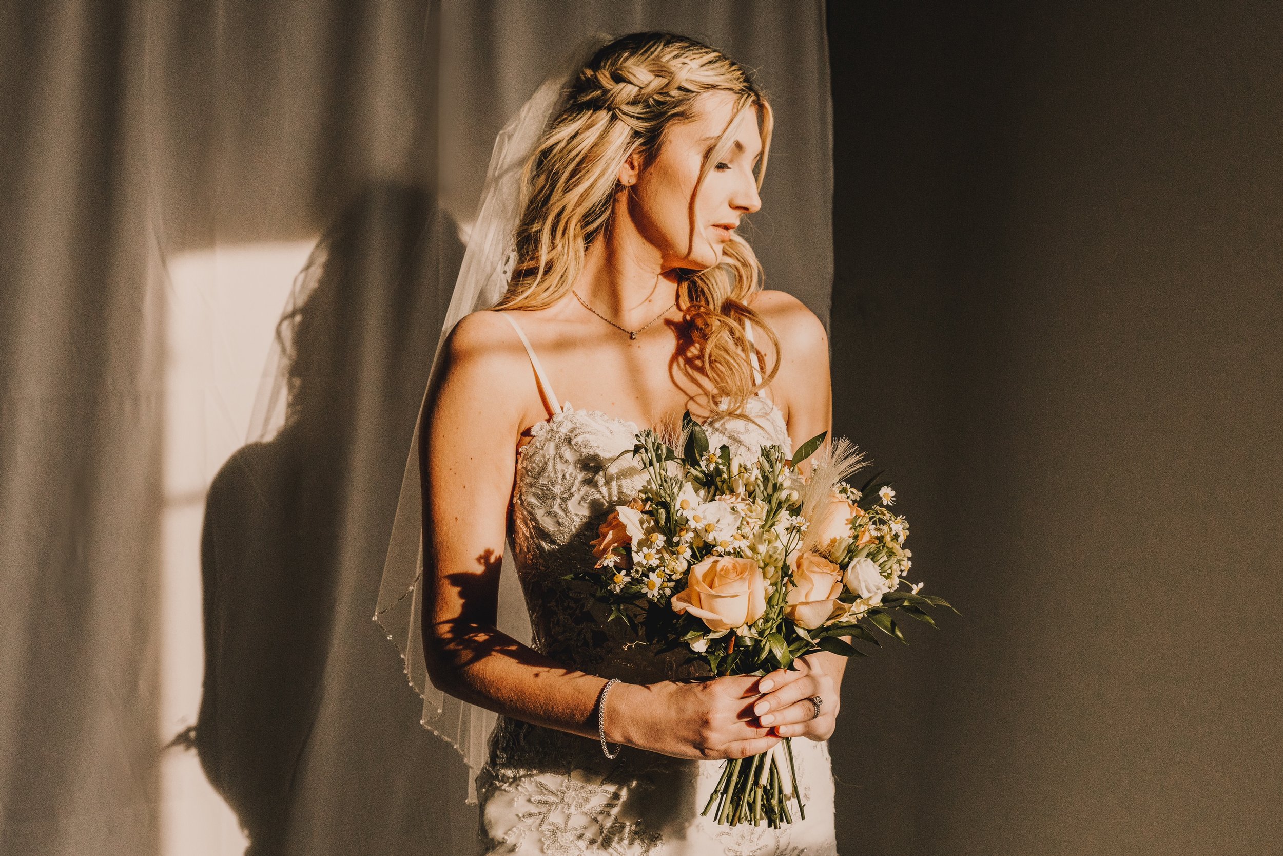 A bride with wavy blonde hair in a white lace wedding dress holding a bouquet of peach roses, white daisies, and greenery, standing against a shadowy background with soft side lighting.