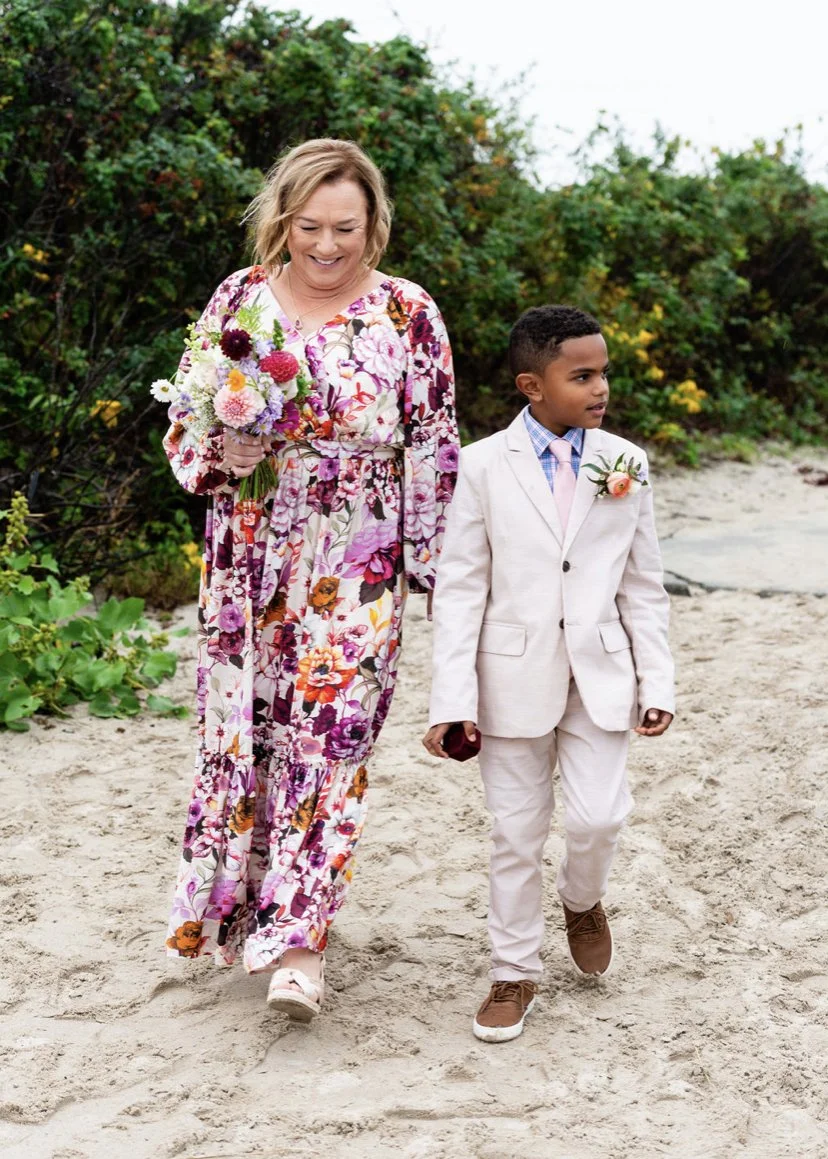 A woman in a long floral dress walking on a sandy beach, holding a colorful bouquet of flowers, beside a young boy in a light-colored suit with a boutonnière, also walking on the beach during daytime.