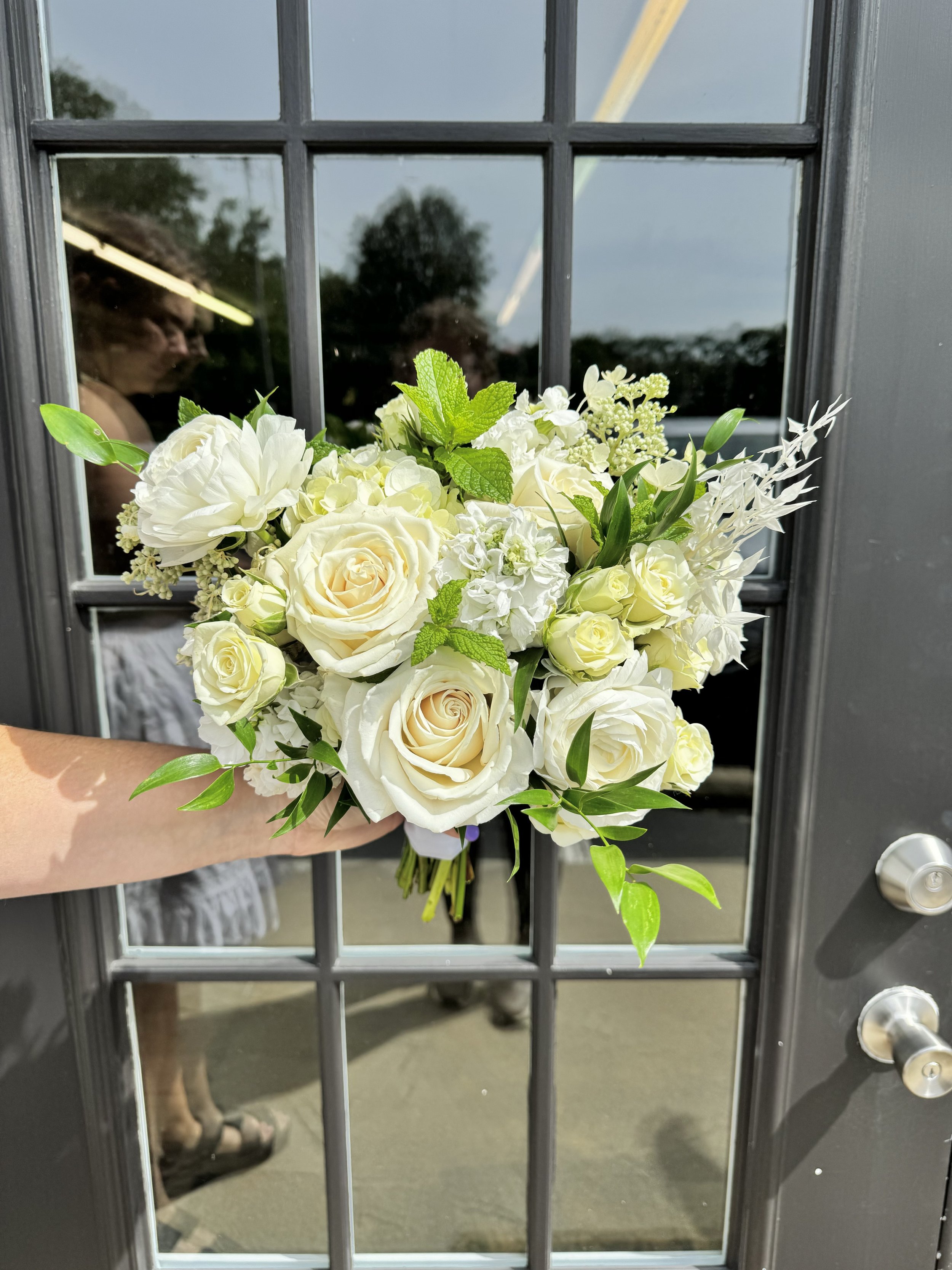 A bouquet of white roses, peonies, and greenery held in front of a glass window on a door, with reflections of people and the outdoors visible in the glass.