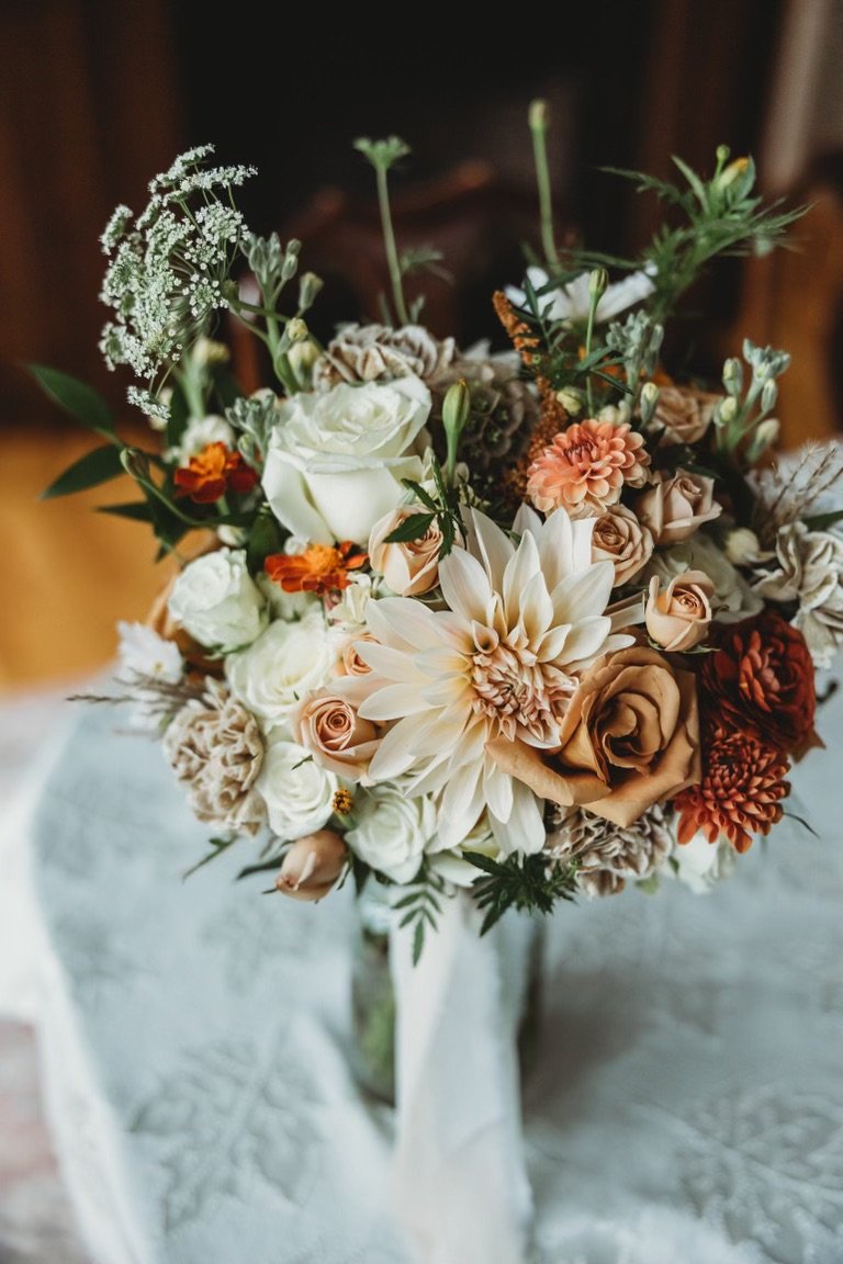 A bouquet of various flowers including roses, dahlias, and greenery in a glass vase on a white cloth surface.