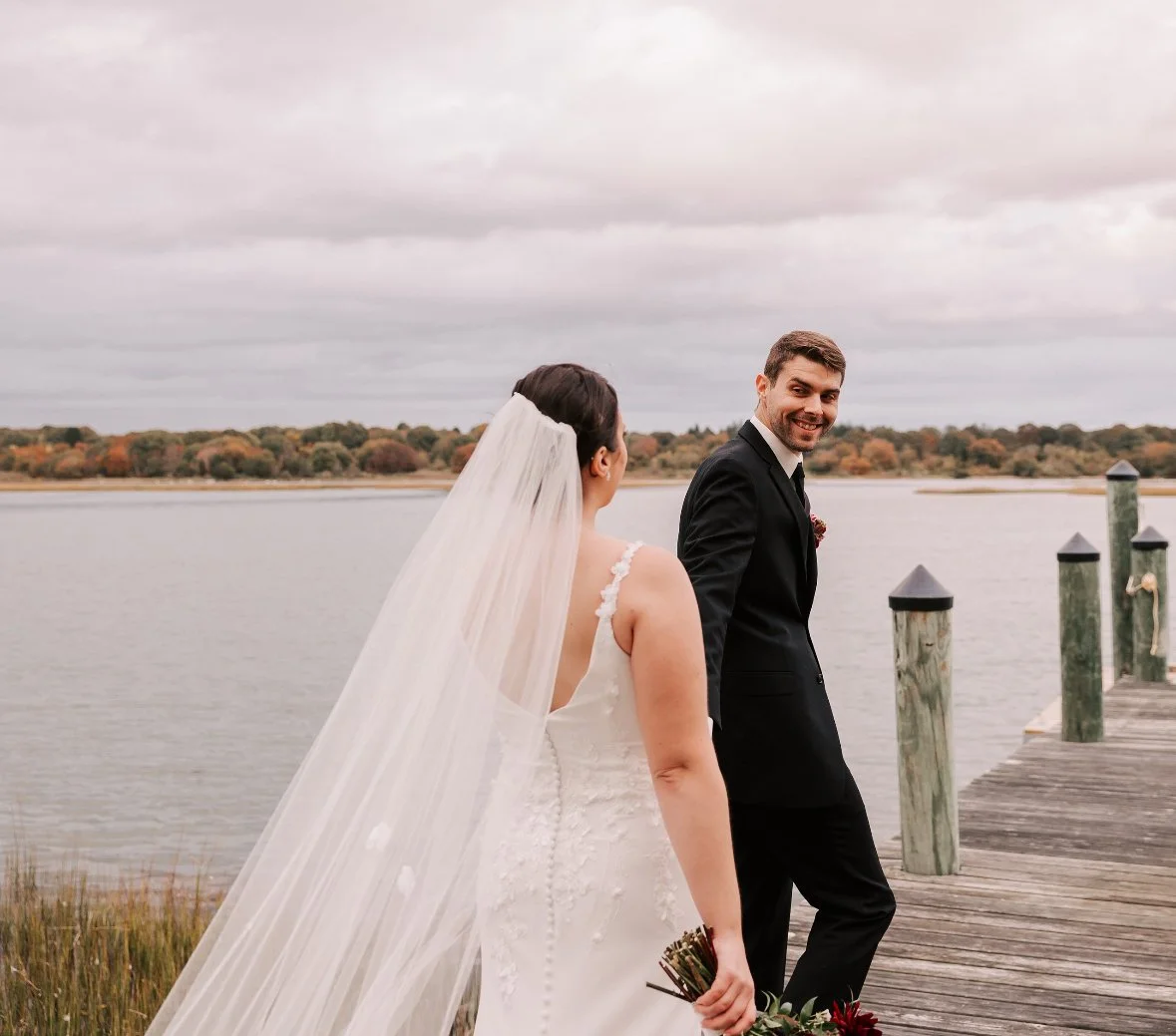 Bride and groom walking on a wooden dock by a body of water, with scenic autumn trees in the background.