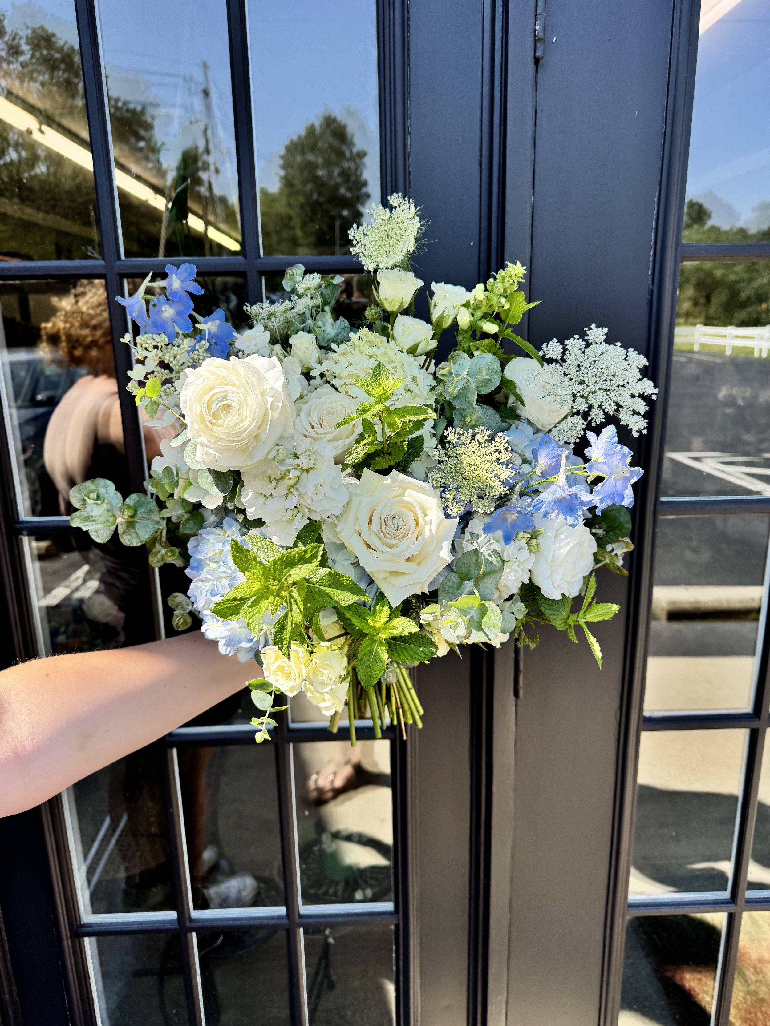 A bouquet of white and light blue flowers held by a person against a black glass door with reflections of trees and people outside.
