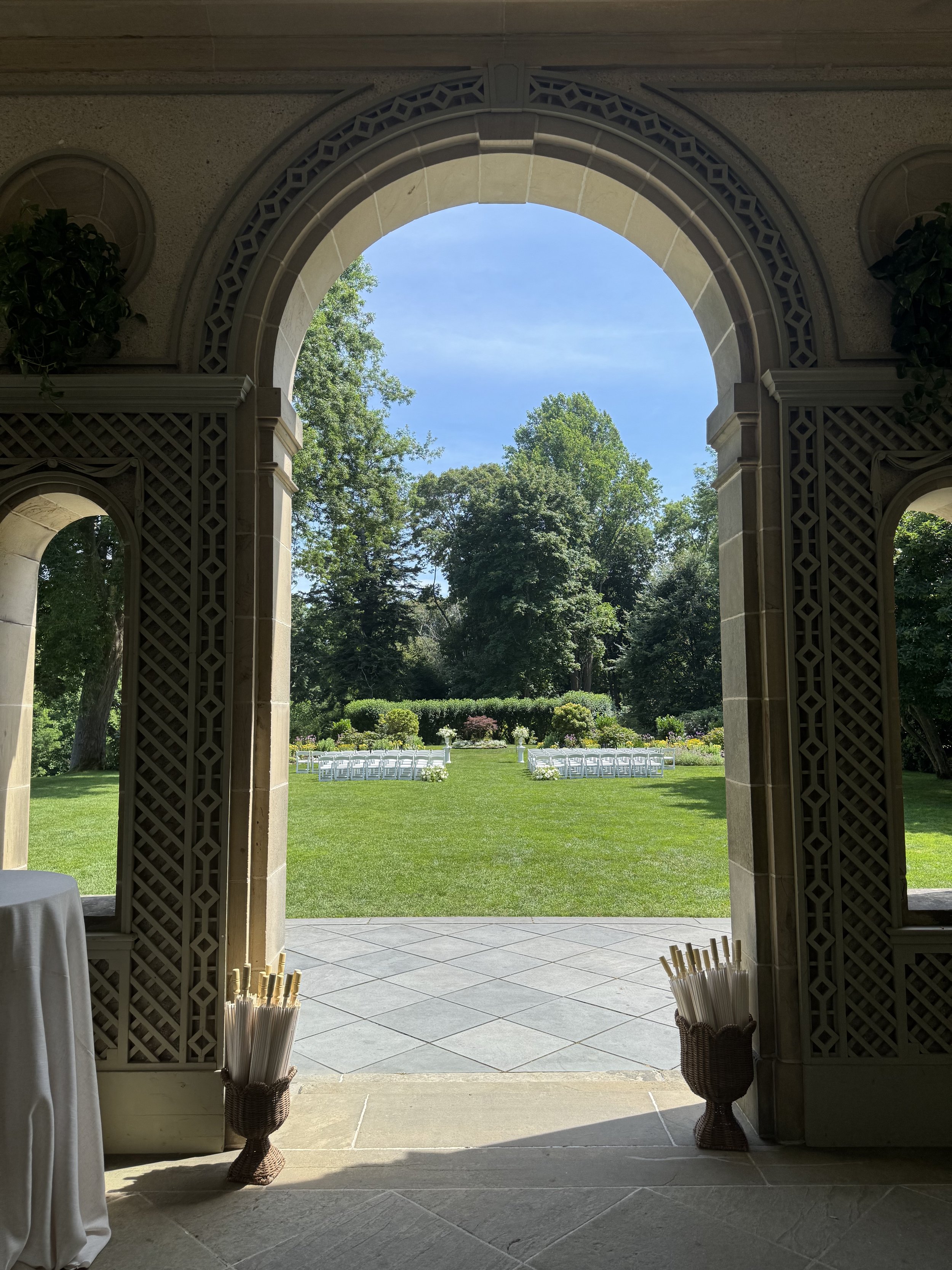 View through a large archway showing a garden with arranged chairs and greenery under a blue sky.