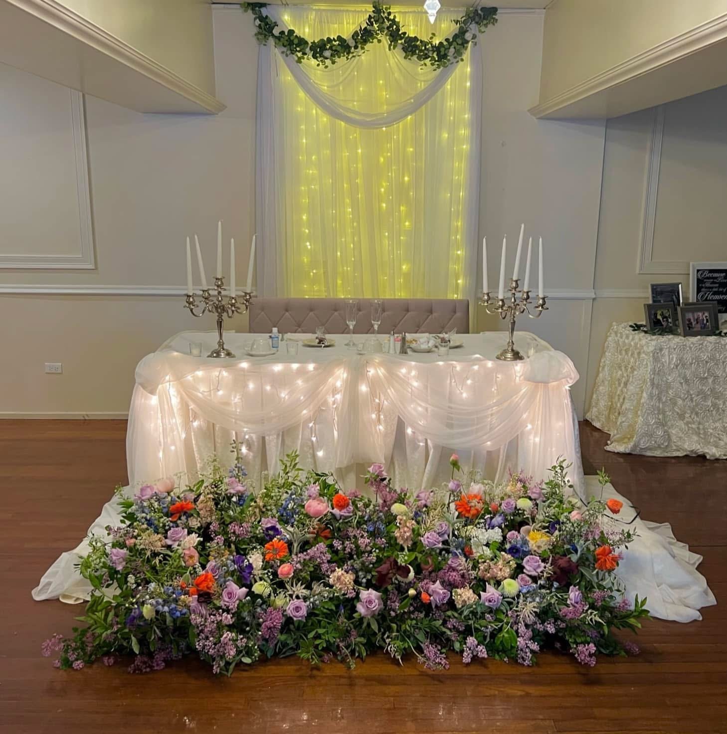 Wedding reception table decorated with white draped fabric, string and candle chandeliers, surrounded by colorful floral arrangements. Behind the table, sheer yellow curtains with fairy lights and greenery garland are hung on the wall.