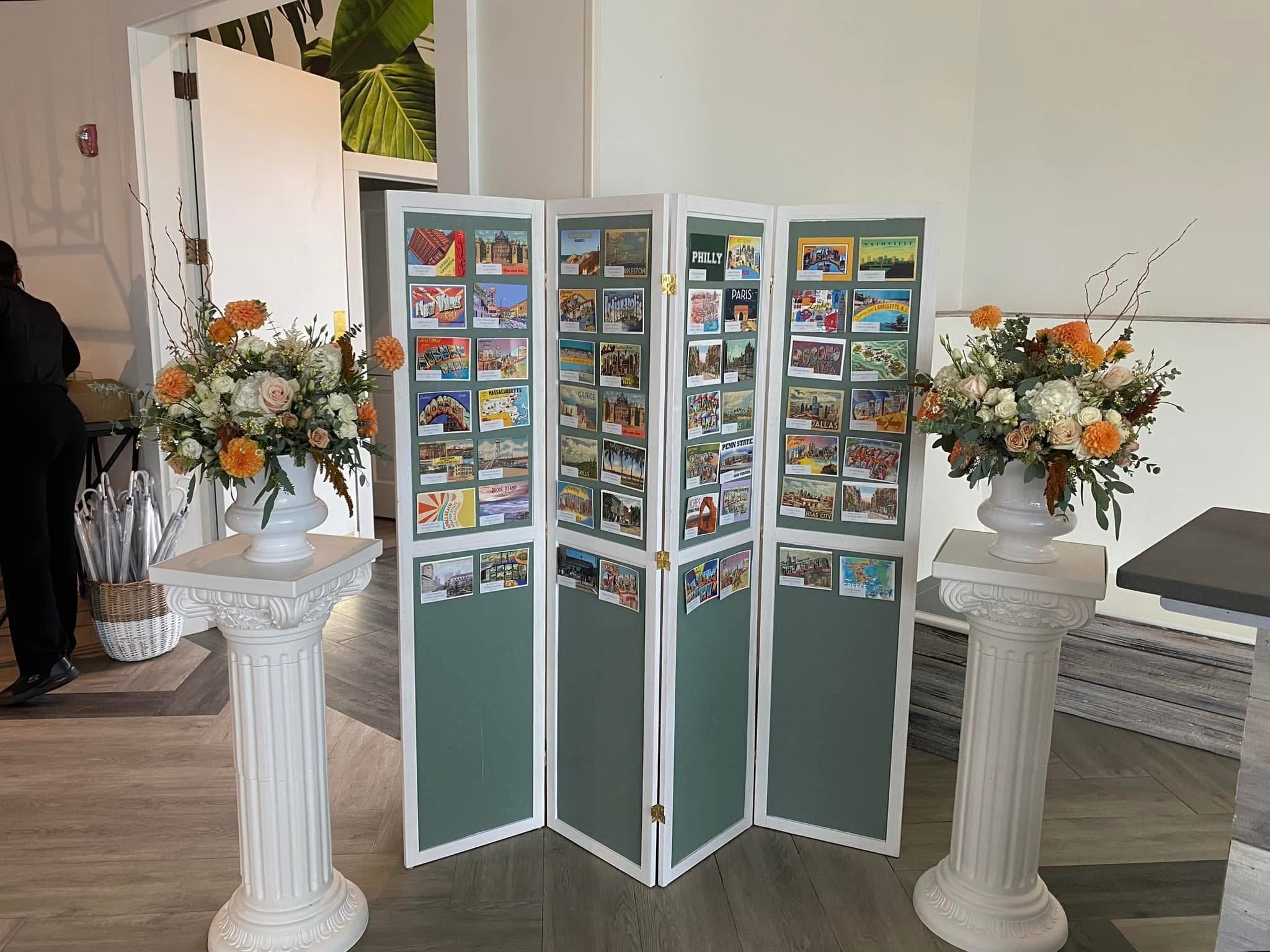 A photo of a display board with various colorful postcards or pictures, flanked by two white columns each topped with floral arrangements, inside a room with a wooden and tiled floor.
