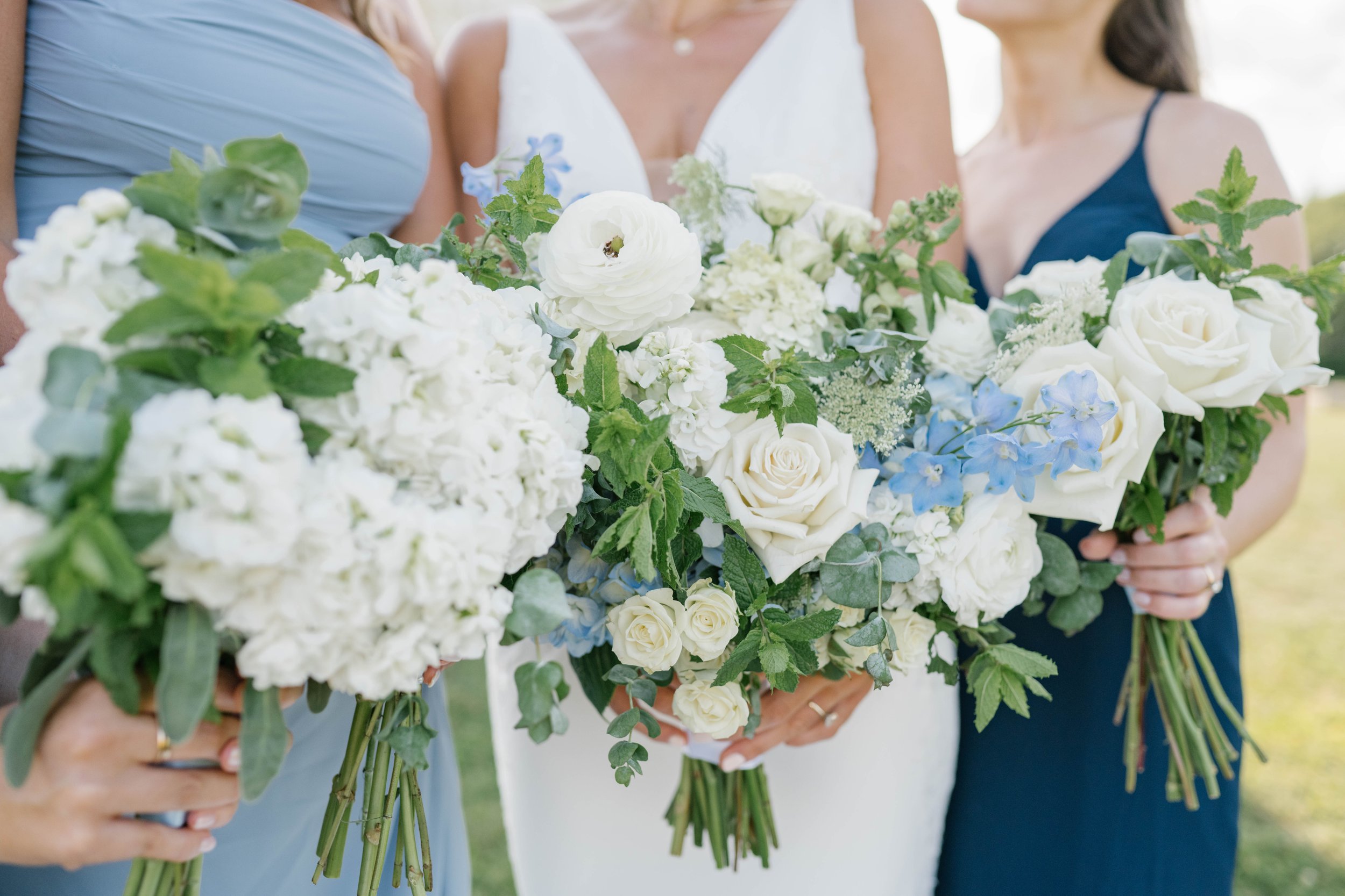 Three women holding large bouquets of white and light blue flowers, including roses and hydrangeas, at an outdoor event or wedding.