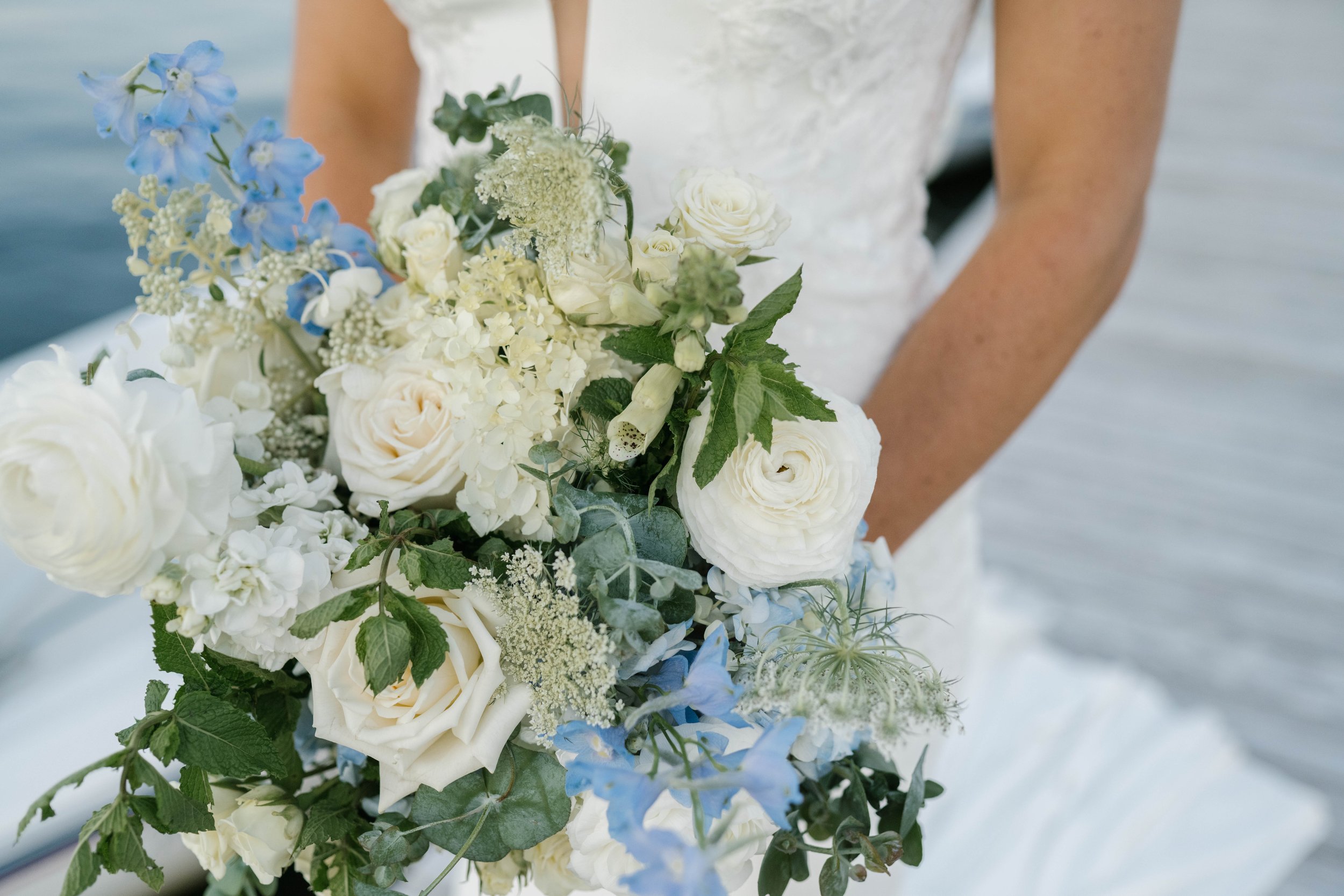 Close-up of a wedding bouquet with white roses, hydrangeas, blue delphiniums, and greenery, held by a bride in a white wedding dress.