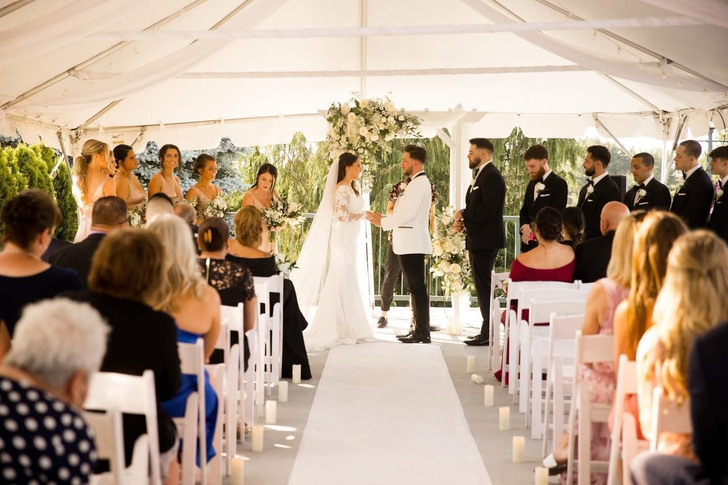A wedding ceremony under a large tent with the bride and groom exchanging vows, surrounded by bridesmaids and groomsmen. Guests seated on either side watch the couple.