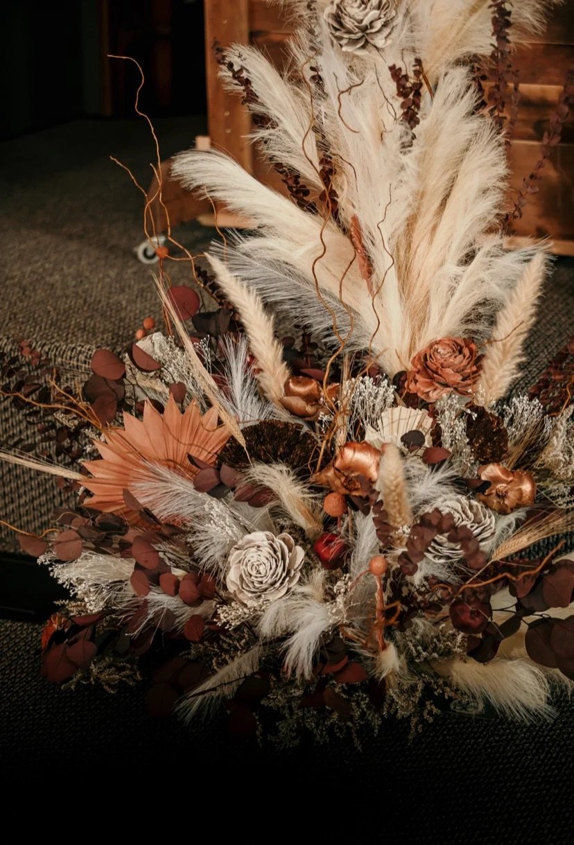 Arrangement of dried flowers and plants with beige, brown, and copper tones, including pampas grass, roses, and various foliage, styled in a rustic setting.