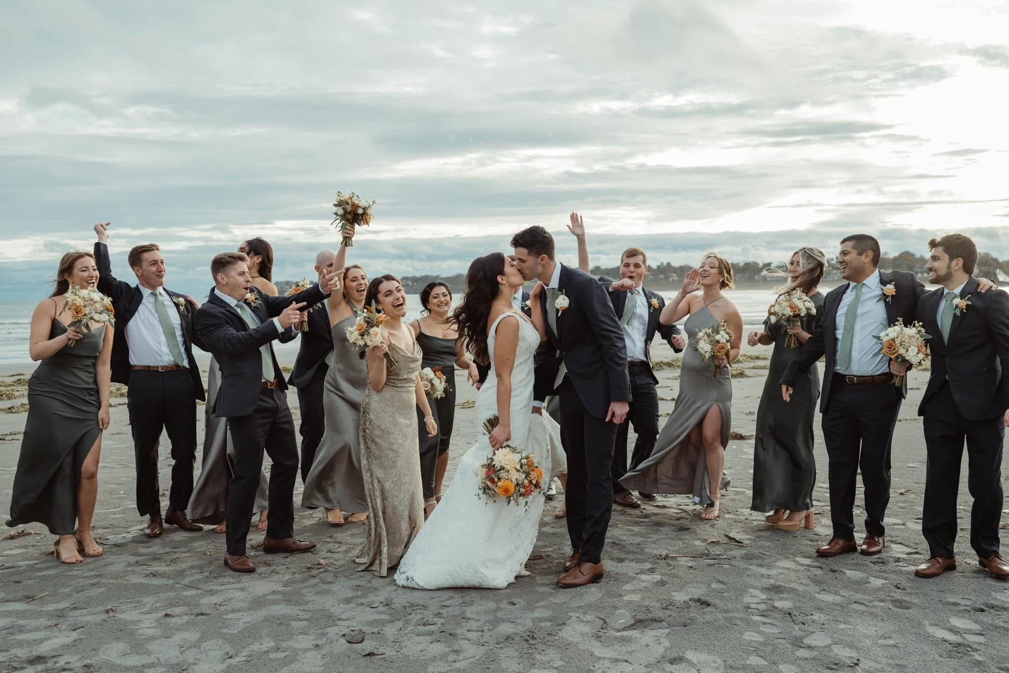 A wedding party on the beach, with the bride and groom kissing in the center surrounded by bridesmaids and groomsmen celebrating.