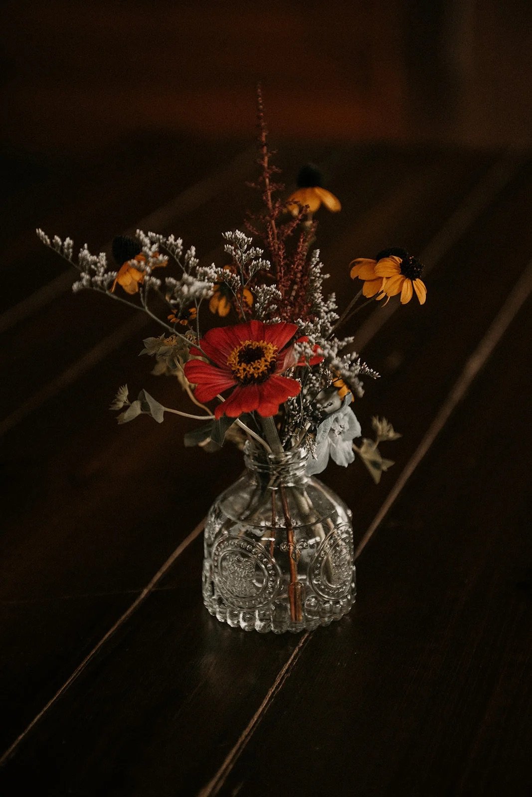 A small glass jar with embossed designs holding a bouquet of mixed dried flowers including yellow, orange, and white blooms, on a dark wooden surface.