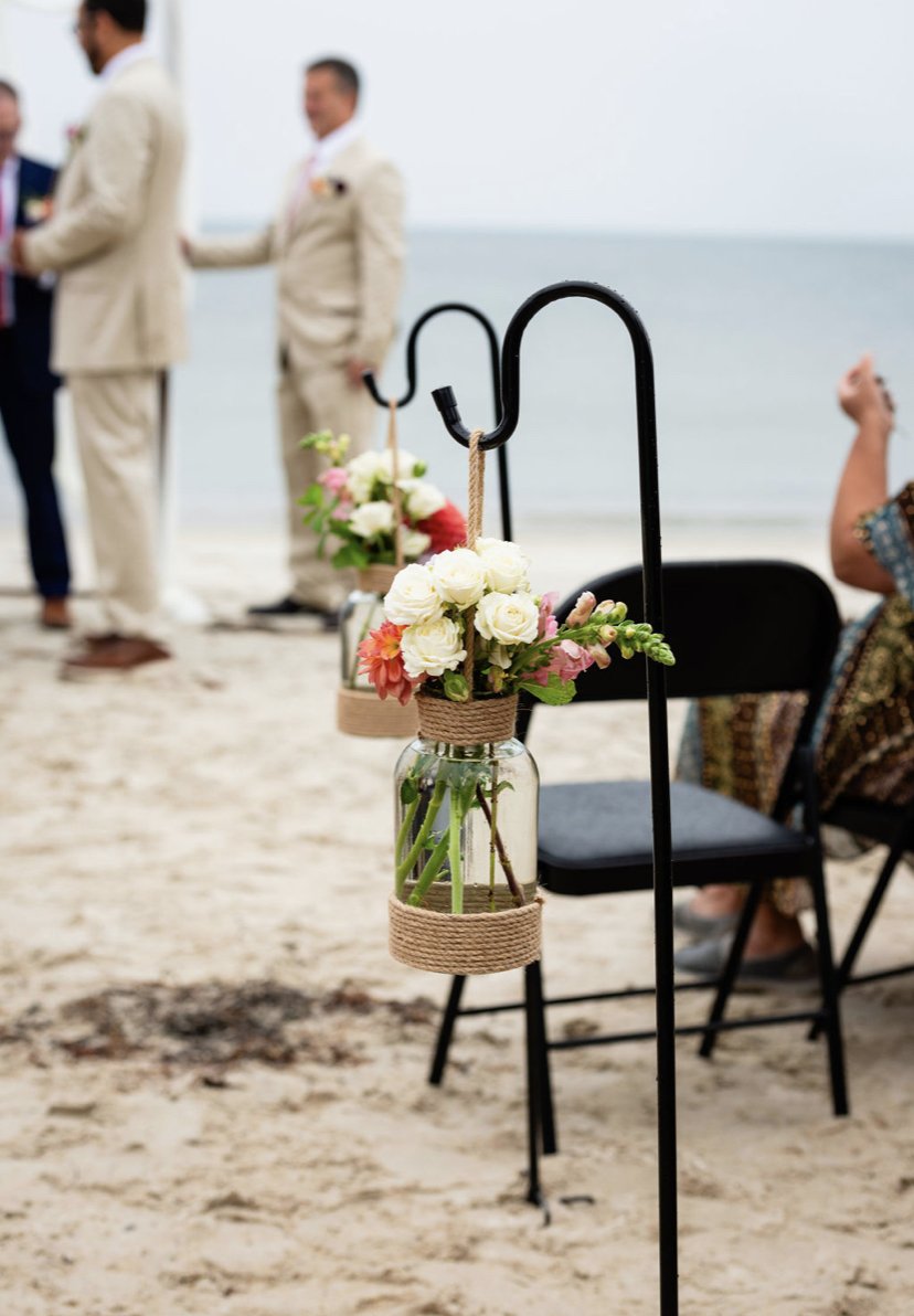 Beach wedding setup with floral decorations hanging from a black metal stand, blurred people in suits and an ocean in the background.