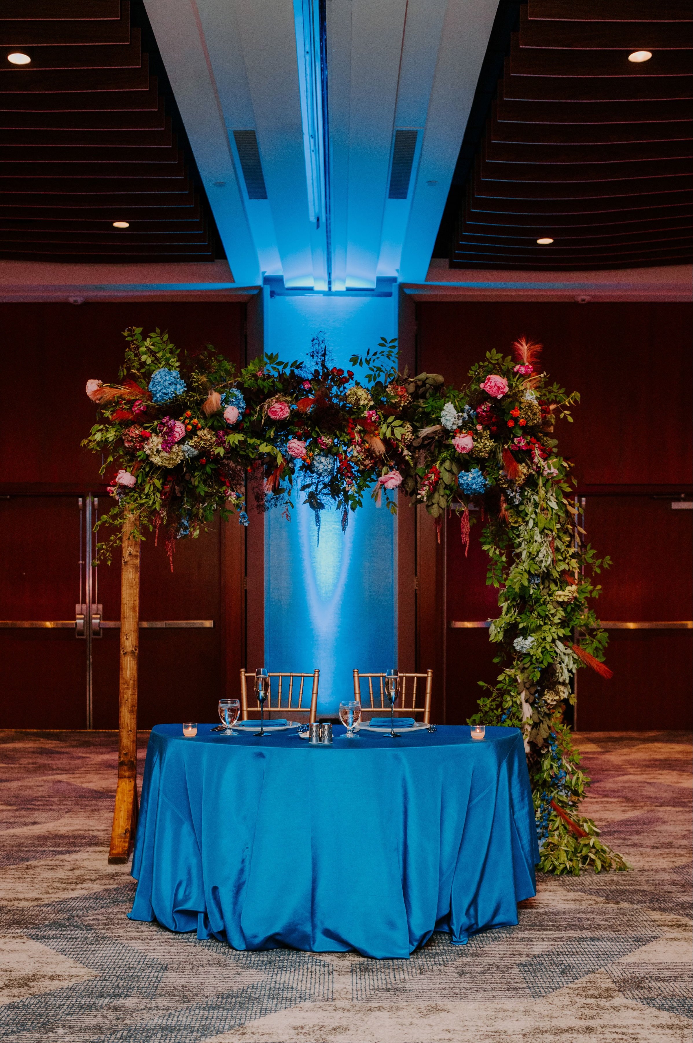 A decorated wedding or event ceremony setup with a round table draped in a blue cloth, two chairs, and a wooden floral arch with colorful flowers and greenery against a blue-lit background in an indoor venue.