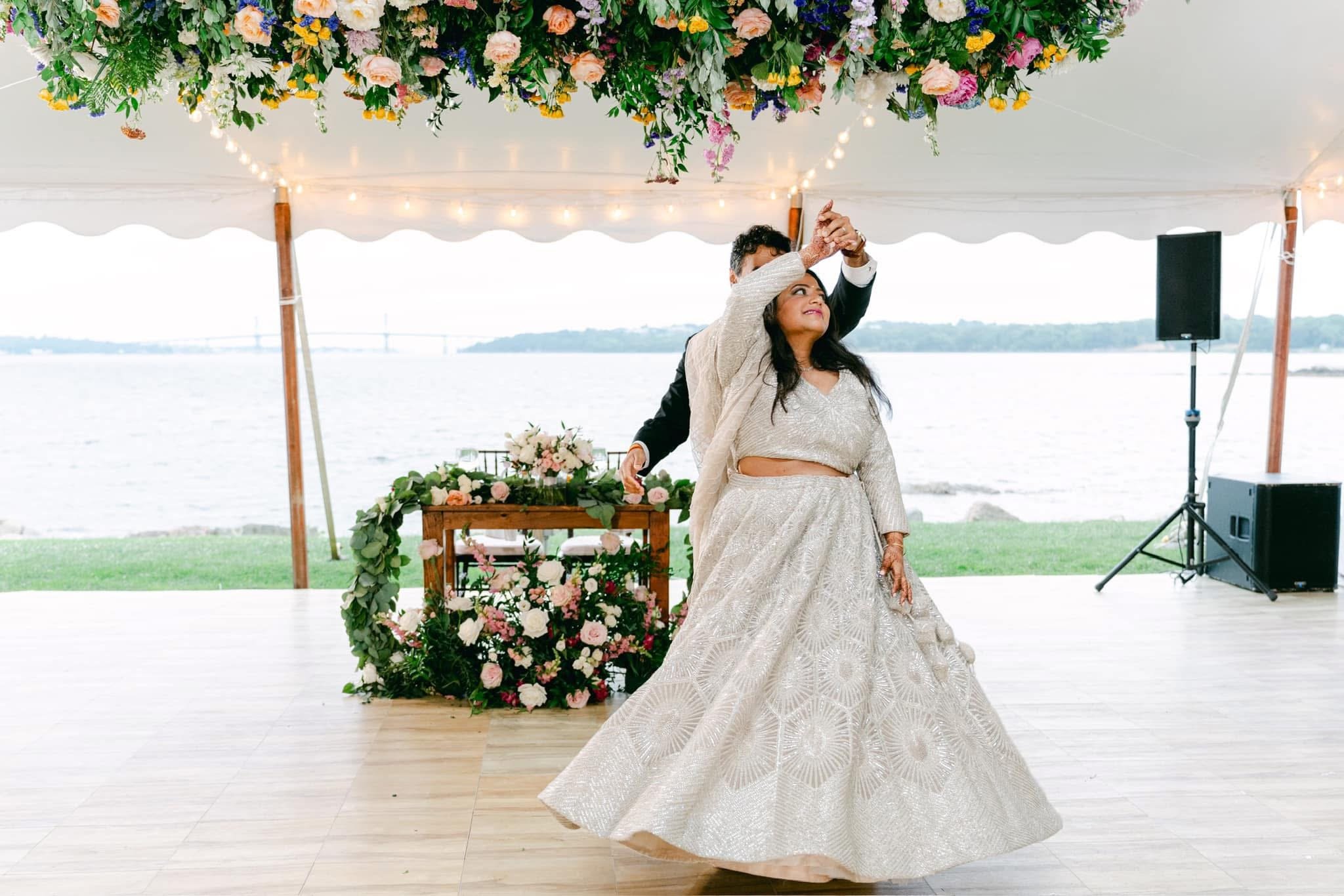 A woman in a traditional wedding dress dances with a man in a suit at an outdoor wedding reception. The ceremony is by a body of water with a bridge in the distance, decorated with flowers and string lights on a tent canopy.