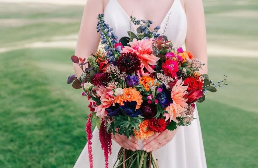 A bride holding a large, colorful bouquet of flowers outdoors on a grassy field.