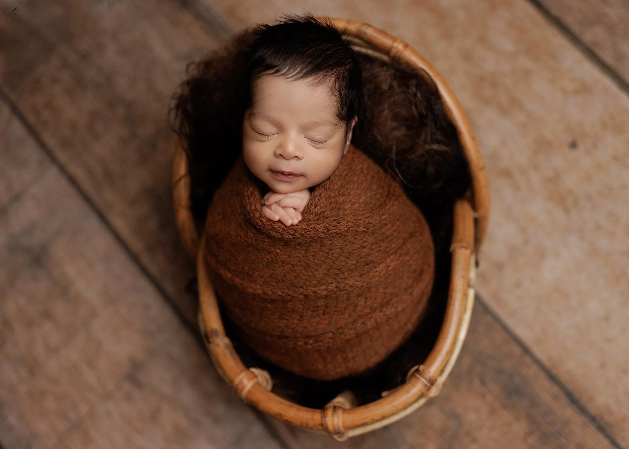 Overhead portrait of a sleeping newborn boy wrapped in rust-brown knit fabric, with hands tucked beneath his chin in an oval woven basket on a wooden backdrop.